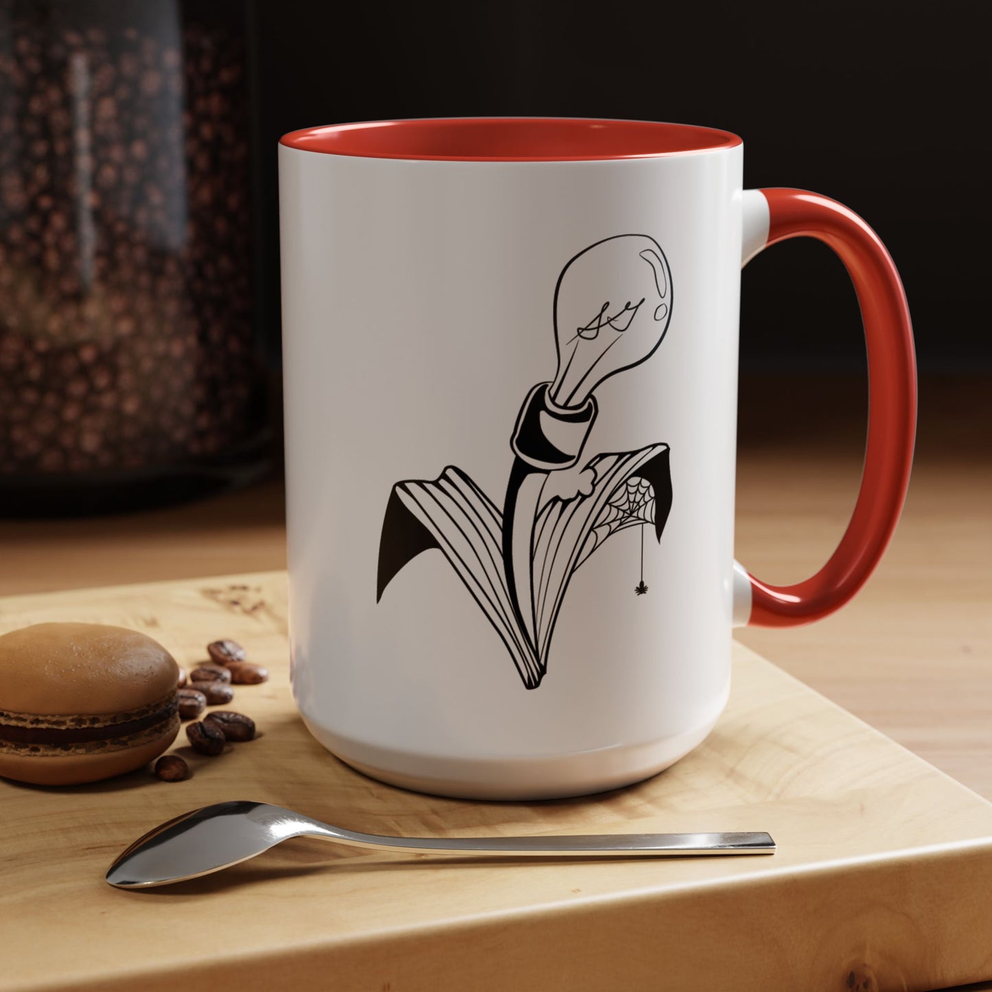 This is the Red 15oz Storm Garden Studio Accent Coffee Mug. This is a mug by Storm Garden Studio which has the logo printed in black and white. The logo is a lightbulb that is screwed onto the leaves of a plant. One leaf is bitten off with a spiderweb. The mug is on top of a light wood block that is on top of a wood desk, a small silver spoon in the front, a chocolate macaroon and some coffee beans to the left. In the background there is a large jar of beans to the left. The wallpaper is black/grey.