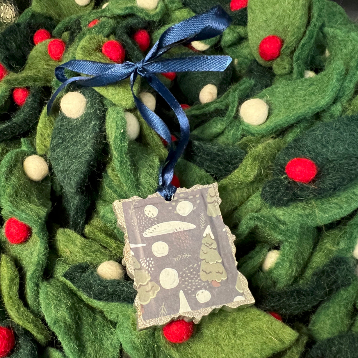 Close-up of snow-laden pine branches and textured orange ribbon on the ornament’s rectangular wooden frame highlighting