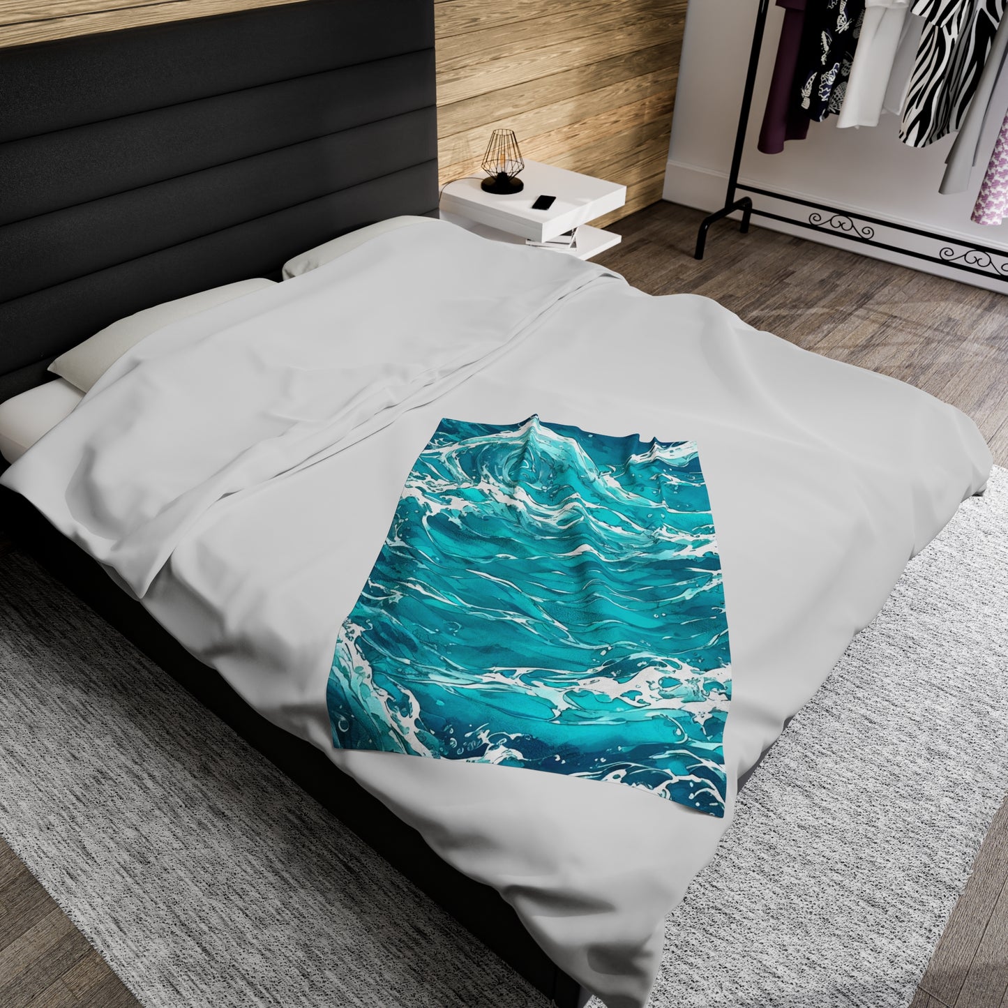 The Swirling Seas blanket by Lee Hansheng Studios is placed centered on a modern queen sized bed dressed with a white duvet, and black upholstered headboard. The blanket features waves in mixed hues of blues and greens, swirling together with foam caps. White nightstand to right of bed with black wire lamp on top. Pine wooden walls cover wall behind bed. A clothing rack stands in distance against a white wall. Flooring is grey brown wood with large light grey rug centered under the bed.