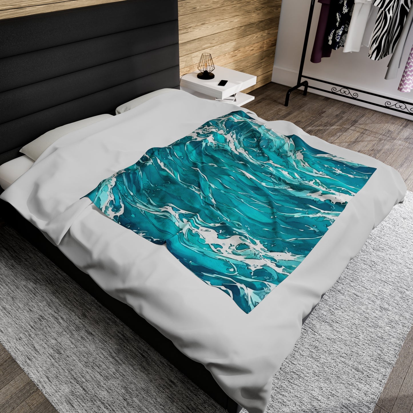 The Swirling Seas blanket by Lee Hansheng Studios is placed centered on a modern queen sized bed dressed with a white duvet, and black upholstered headboard. The blanket features waves in mixed hues of blues and greens, swirling together with foam caps. White nightstand to right of bed with black wire lamp on top. Pine wooden walls cover wall behind bed. A clothing rack stands in distance against a white wall. Flooring is grey brown wood with large light grey rug centered under the bed.