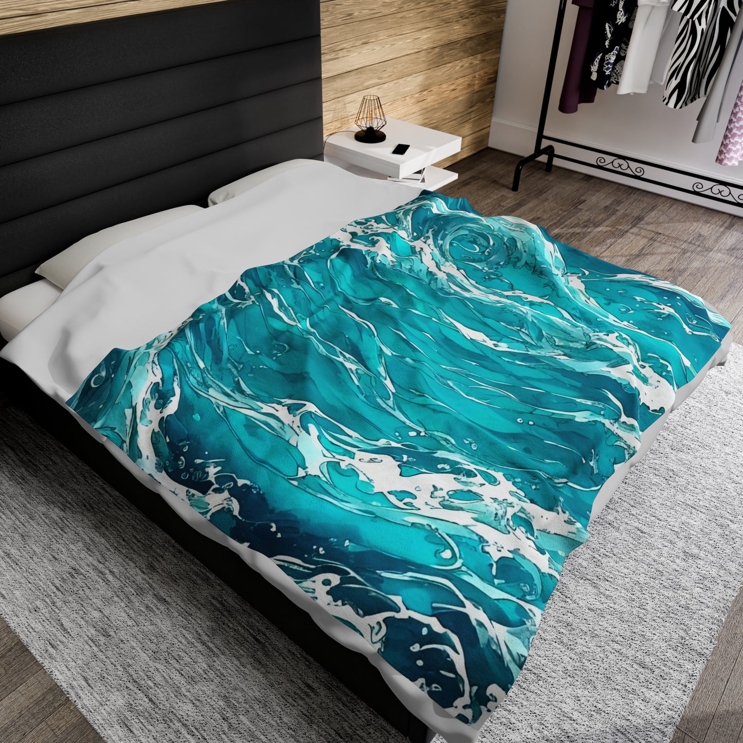 The Swirling Seas design by Lee Hansheng Studios is placed centered on a modern bed made with a white duvet, and black upholstered headboard. the blanket features beautiful waves, in hues of blues and greens with white foam caps. Pine accent wall behind bed. whit wall in distance with a clothes rack in front of it. Rustic grey brown floors with large light grey accent rug beneath bed.