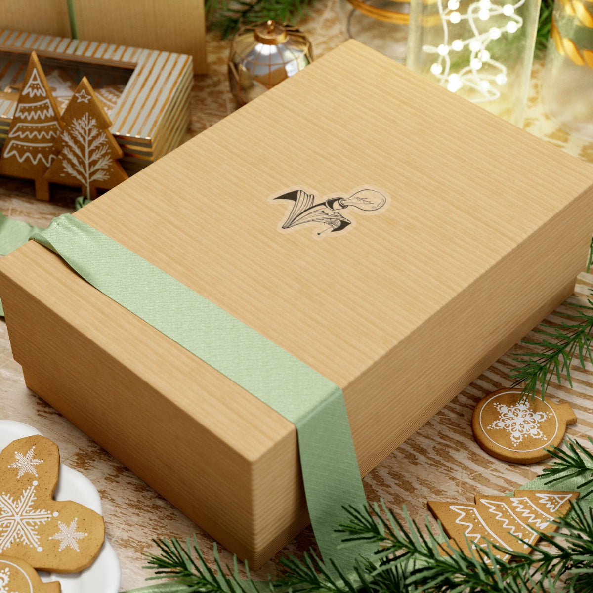 This is the Transparent 2" x 2" Storm Garden Studio Sticker. The sticker is a lightbulb with two leaves. The right leaf is bitten through with a spiderweb and a spider. The sticker is placed on rectangular box with a light green ribbon on the bottom. The box is on top of striped surface. It is surrounded by cardboard christmas decor like trees, snowflakes and ornaments. There is a jar of lights on the top right and pine tree leaves on the top and bottom.