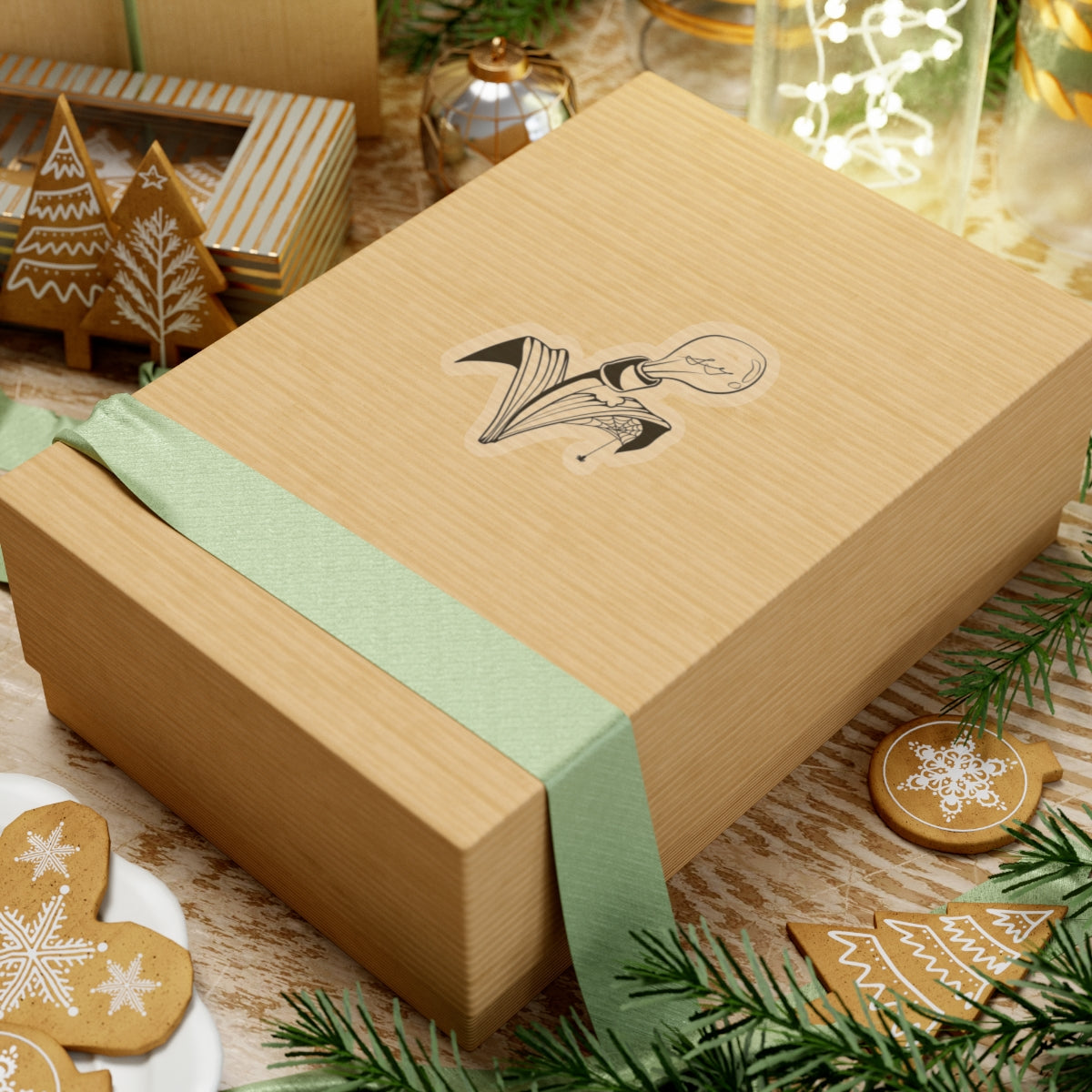 This is the Transparent 4" x 4" Storm Garden Studio Sticker. The sticker is a lightbulb with two leaves. The right leaf is bitten through with a spiderweb and a spider. The sticker is placed on rectangular box with a light green ribbon on the bottom. The box is on top of striped surface. It is surrounded by cardboard christmas decor like trees, snowflakes and ornaments. There is a jar of lights on the top right and pine tree leaves on the top and bottom.