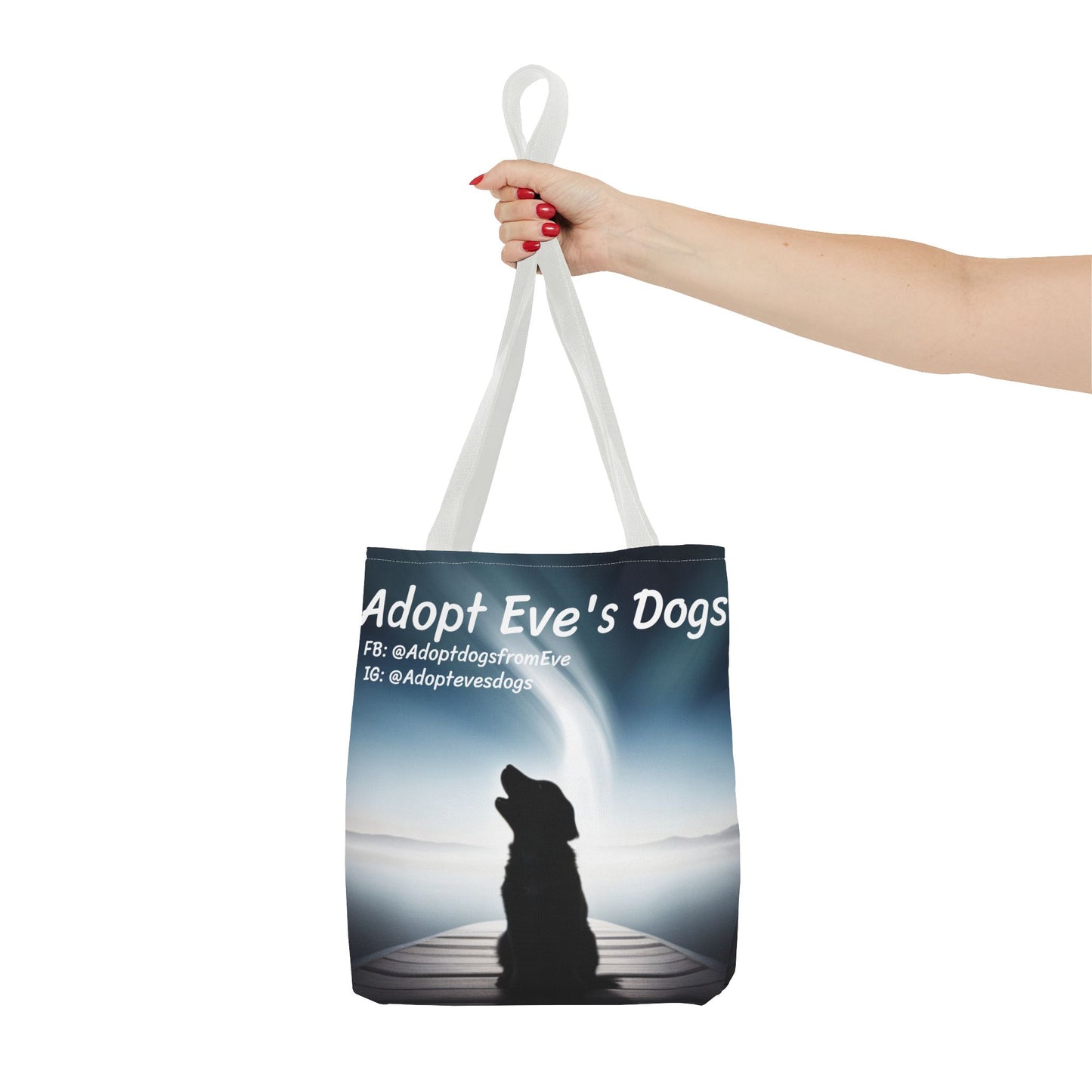 This is the White 13" x 13" Adopt Eve's Dogs Tote with color matched handles. This is an all around print tote with "Adopt Eve's Dogs" and socials on the top in white. The print is of a shadow of a golden retriever looking to the left on a pier towards water with mountains surrounding it. The image is shown with the upper arm to the hand. The hand holding the handles of tote from the right with red nail polish. The tote is against a white background.