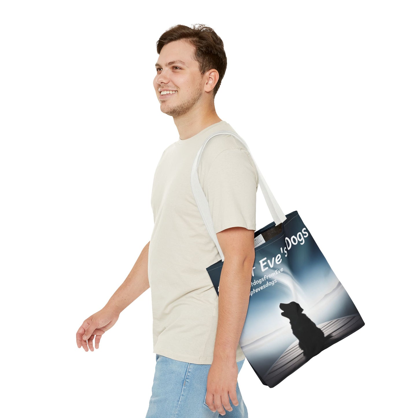 This is the White 16" x 16" Adopt Eve's Dogs Tote with color matched handles. This is an all around print tote with "Adopt Eve's Dogs" and socials on the top in white. The print is of a shadow of a golden retriever looking to the left on a pier towards water with mountains surrounding it. There is a smiling brunette man walking to the left, looking to the left, with the tote over his shoulder. He is wearing a white tee and light jeans. He is against a white background.