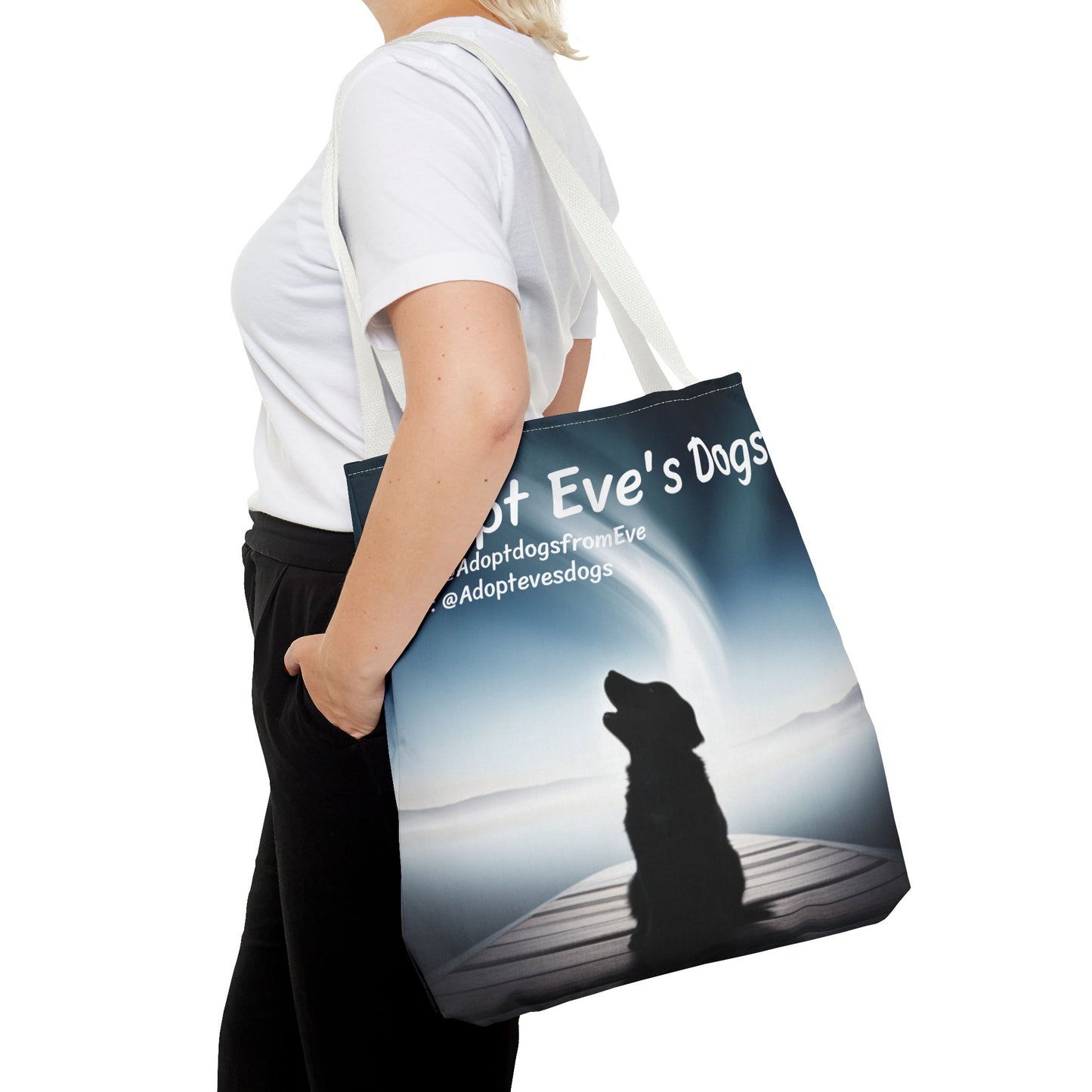 This is the White 18" x 18" Adopt Eve's Dogs Tote with color matched handles. This is an all around print tote with "Adopt Eve's Dogs" and socials on the top in white. The print is of a shadow of a golden retriever looking to the left on a pier towards water with mountains surrounding it. There is a woman wearing a white tee, black pants and short blond hair. Her hand is in the pants pocket and has the tote over her shoulder. She is against a white background.