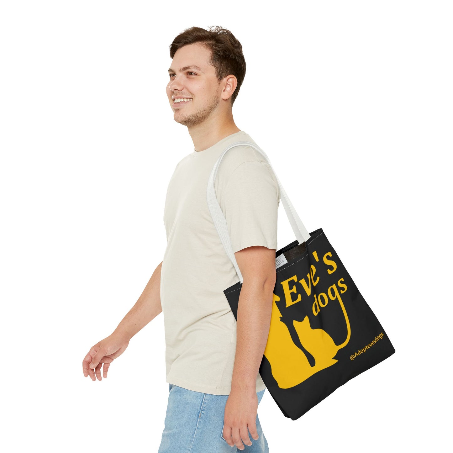 This is the White 13" x 13" Adopt Eve's Dog Tote Bag. The handles are white. The top of the tote is hemmed in yellow. The tote is all over black with the image of a dog, cat, "Eve's Dogs" and "@Adoptevesdogs" in yellow. A man with a smile, short wavy brown hair, beige tee plus jeans with tote around their shoulder and walking towards the left against a white background.
