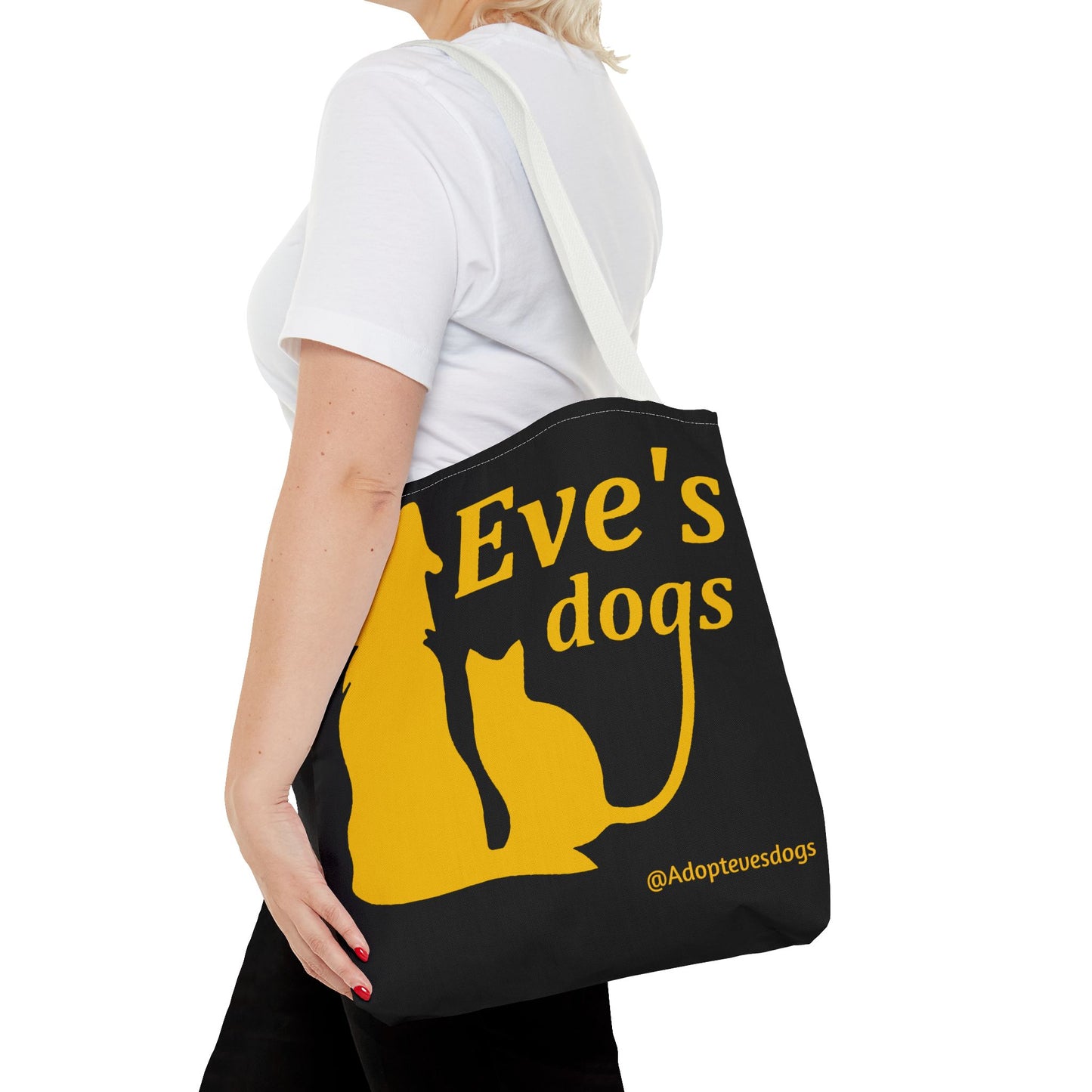This is the White 16" x 16" Adopt Eve's Dog Tote Bag. The handles are white. The tote is all over black with the image of a dog, cat, "Eve's Dogs" and "@Adoptevesdogs" in yellow. It is over the shoulder of a short blonde haired woman wearing a white tea and black pants. Her hand with red nails is on the tote and she is facing the left.