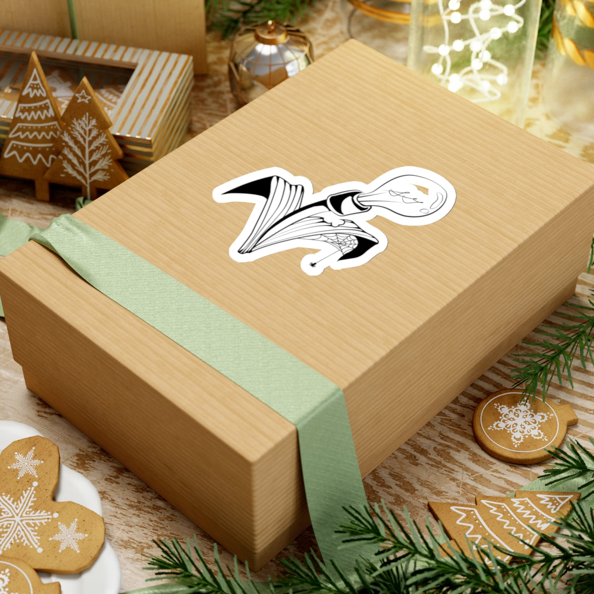 This is the White 4" x 4" Storm Garden Studio Sticker. The sticker is a lightbulb with two leaves. The right leaf is bitten through with a spiderweb and a spider. The sticker is placed on rectangular box with a light green ribbon on the bottom. The box is on top of striped surface. It is surrounded by cardboard christmas decor like trees, snowflakes and ornaments. There is a jar of lights on the top right and pine tree leaves on the top and bottom.