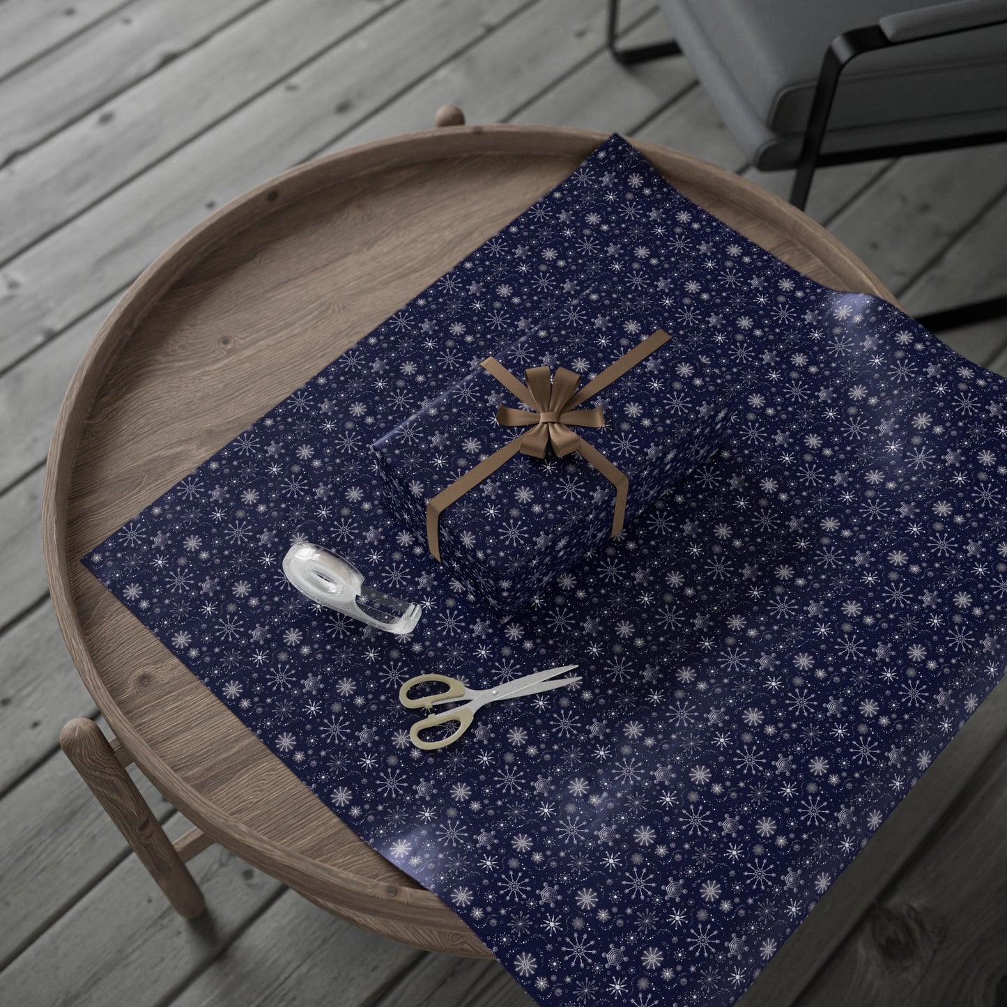 This is the glossy 30" x 36" Winter Blizzard Gift Wrap. It is placed on a small dark brown wood coffee side table. On top is the gift wrap roll on the left and below is a sheet that was cut out with a rectangular gift wrapped slightly to the right. A brown string covers each side and tied in a bow in the middle. Below the gift is small tan scissors and clear tape roll. The background is a gray wood flooring and in the upper right is a faux leather gray chair.