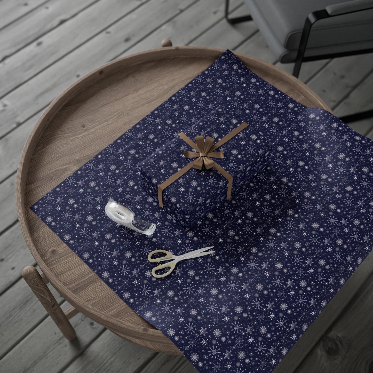 This is the glossy 30" x 72" Winter Blizzard Gift Wrap. It is placed on a small dark brown wood coffee side table. On top is the gift wrap roll on the left and below is a sheet that was cut out with a rectangular gift wrapped slightly to the right. A brown string covers each side and tied in a bow in the middle. Below the gift is small tan scissors and clear tape roll. The background is a gray wood flooring and in the upper right is a faux leather gray chair.