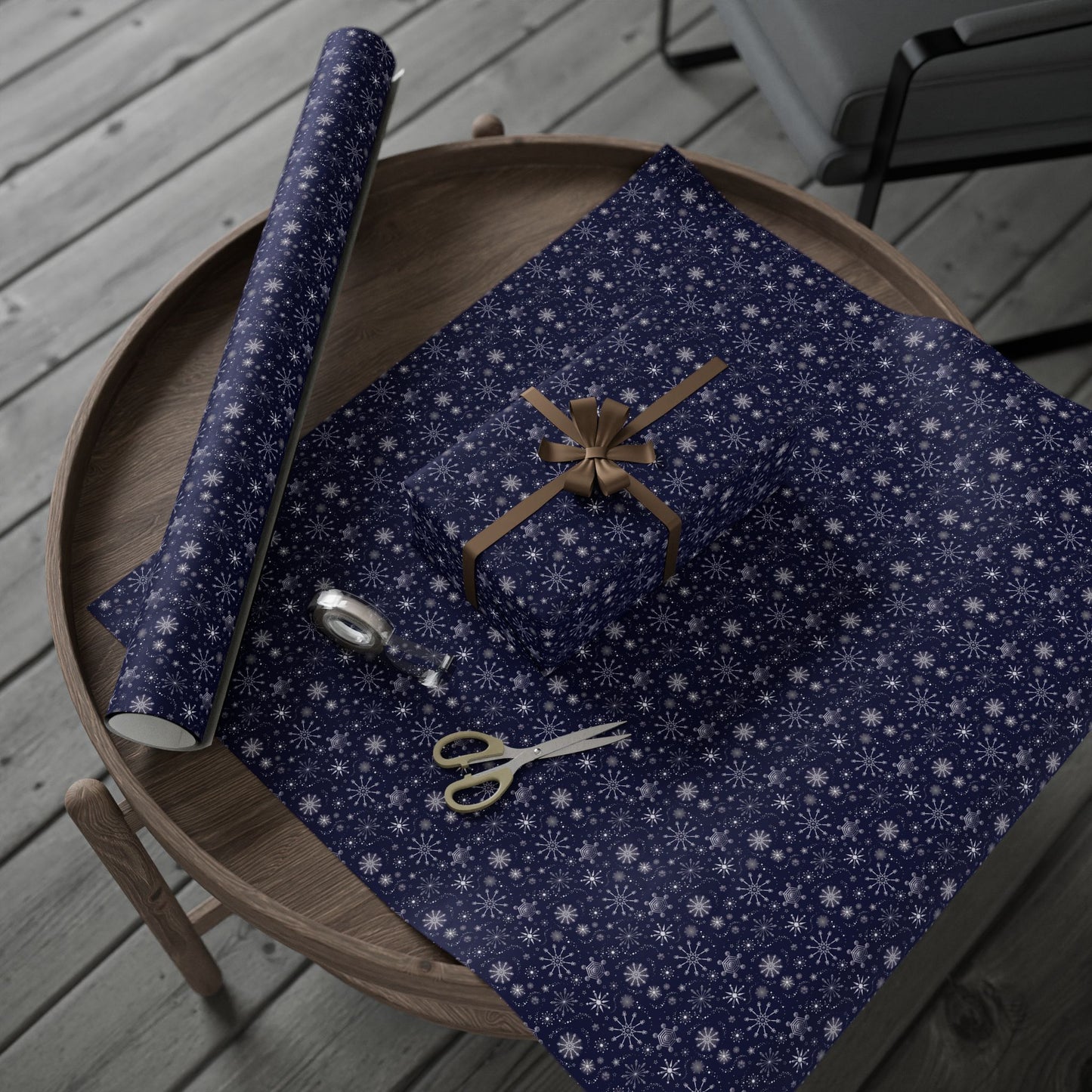 This is the matte 30" x 180" Winter Blizzard Gift Wrap. It is placed on a small dark brown wood coffee side table. On top is the gift wrap roll on the left and below is a sheet that was cut out with a rectangular gift wrapped slightly to the right. A brown string covers each side and tied in a bow in the middle. Below the gift is small tan scissors and clear tape roll. The background is a gray wood flooring and in the upper right is a faux leather gray chair.
