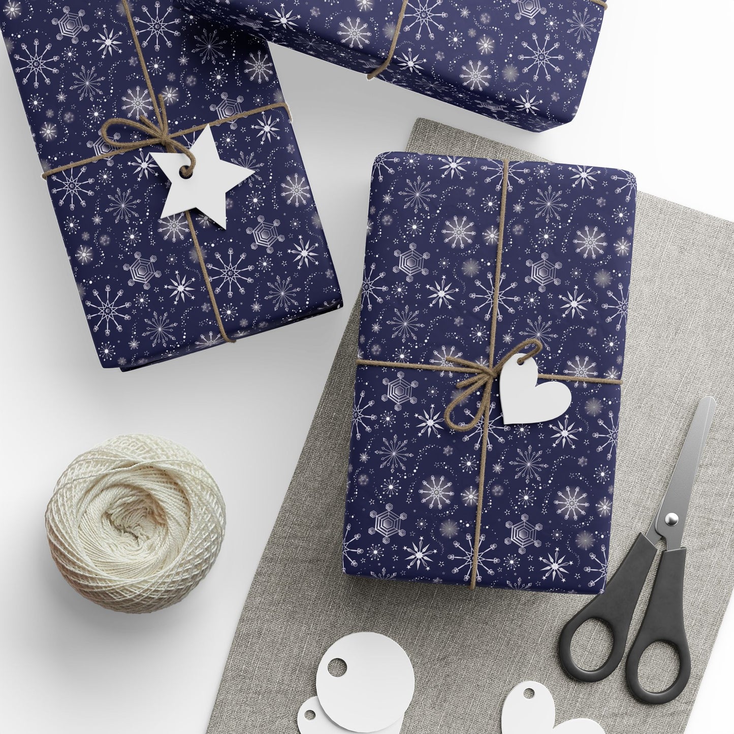 This is the matte 30" x 36" Winter Blizzard Gift Wrap. There are three presents wrapped one slightly on top of the other on the far left all on a white surface. The other is closer to the middle and slight to the right. All gifts are tied with a brown bow. There is a heart and star gift tag on the two gifts that can be seen fully. Below the right one is a grey heather rectangular strip. There are black scissors, circle, and heart tags below. On the left is a role of white thread.