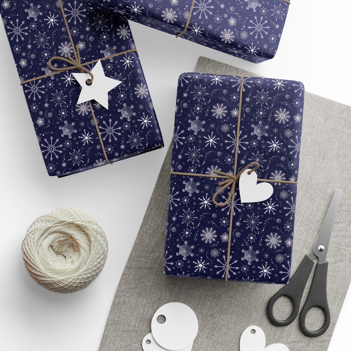 This is the matte 30" x 72" Winter Blizzard Gift Wrap. There are three presents wrapped one slightly on top of the other on the far left all on a white surface. The other is closer to the middle and slight to the right. All gifts are tied with a brown bow. There is a heart and star gift tag on the two gifts that can be seen fully. Below the right one is a grey heather rectangular strip. There are black scissors, circle, and heart tags below. On the left is a role of white thread.