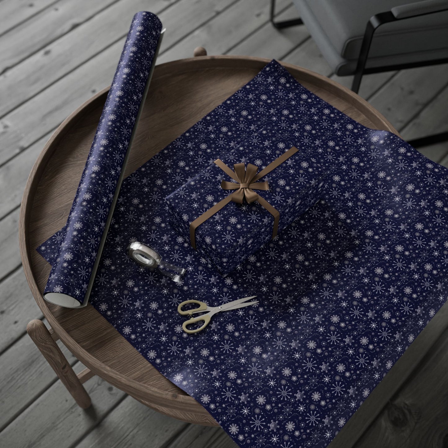 This is the 30" x 72" Winter Blizzard Gift Wrap. It is placed on a small dark brown wood coffee side table. On top is the gift wrap roll on the left and below is a sheet that was cut out with a rectangular gift wrapped slightly to the right. A brown string covers each side and tied in a bow in the middle. Below the gift is small tan scissors and clear tape roll. The background is a gray wood flooring and in the upper right is a faux leather gray chair.