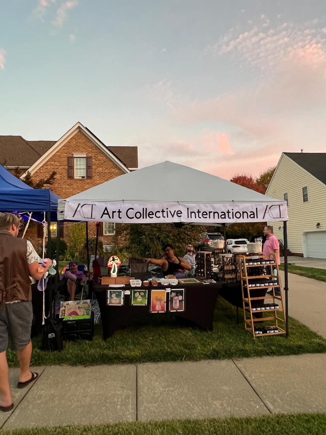 Art Collective International's Tent Set up, white top and blue top tents next to each other, a display of candles on the right, prints in the front, necklaces and pins on turn table displays to the right. 