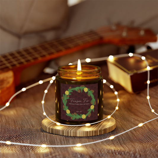 This is the 4oz Amber Fraser Fir Candle from Chris Foster Design. This is a photo from the front open and the wick lit. The candle is in a glass cylinder container that is tinted amber. The label is brown with a delicate pattern and has a lemon and pinecone wreath surround the name of the candle. The candle is on a wood coaster surrounded by little lights. It is on a clothed surface and in the background there is a light wooded ukulele on the left and a brown book on the right and a tan leather sofa.