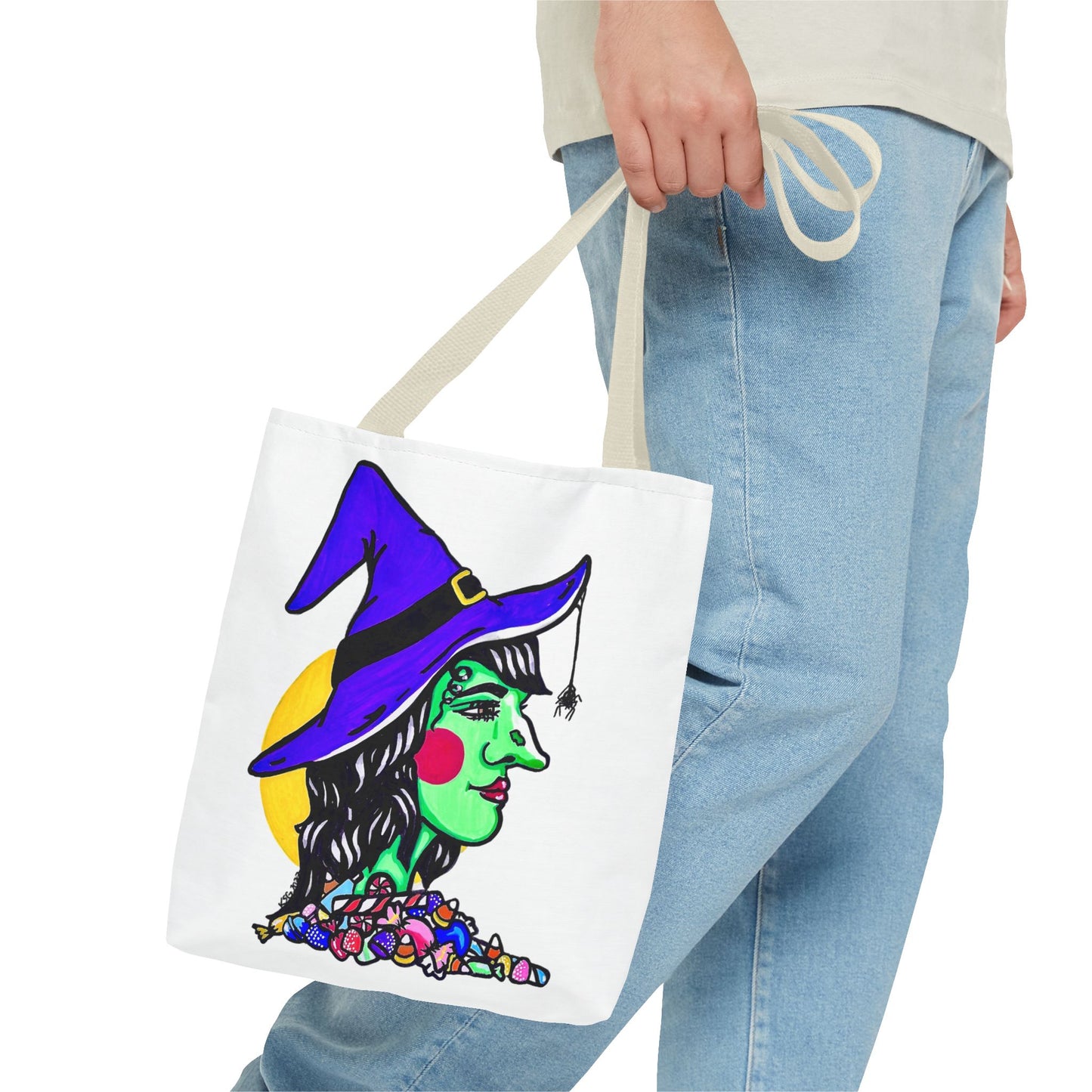 This is the Beige 13" x 13" Witch Tote Bag by Storm Garden Studio. The handles are color matched. It shows the side profile of a witch. She has a purple witch hat, a black belt on hat, yellow buckle, a spider crawling down the front, red rosy cheeks, red lips and assorted candy under the neck. Behind her is a bright yellow circle past her hair. A man's hand with a beige tee and light- colored jeans is grabbing the handles from the hip down. The tote is against a white background.