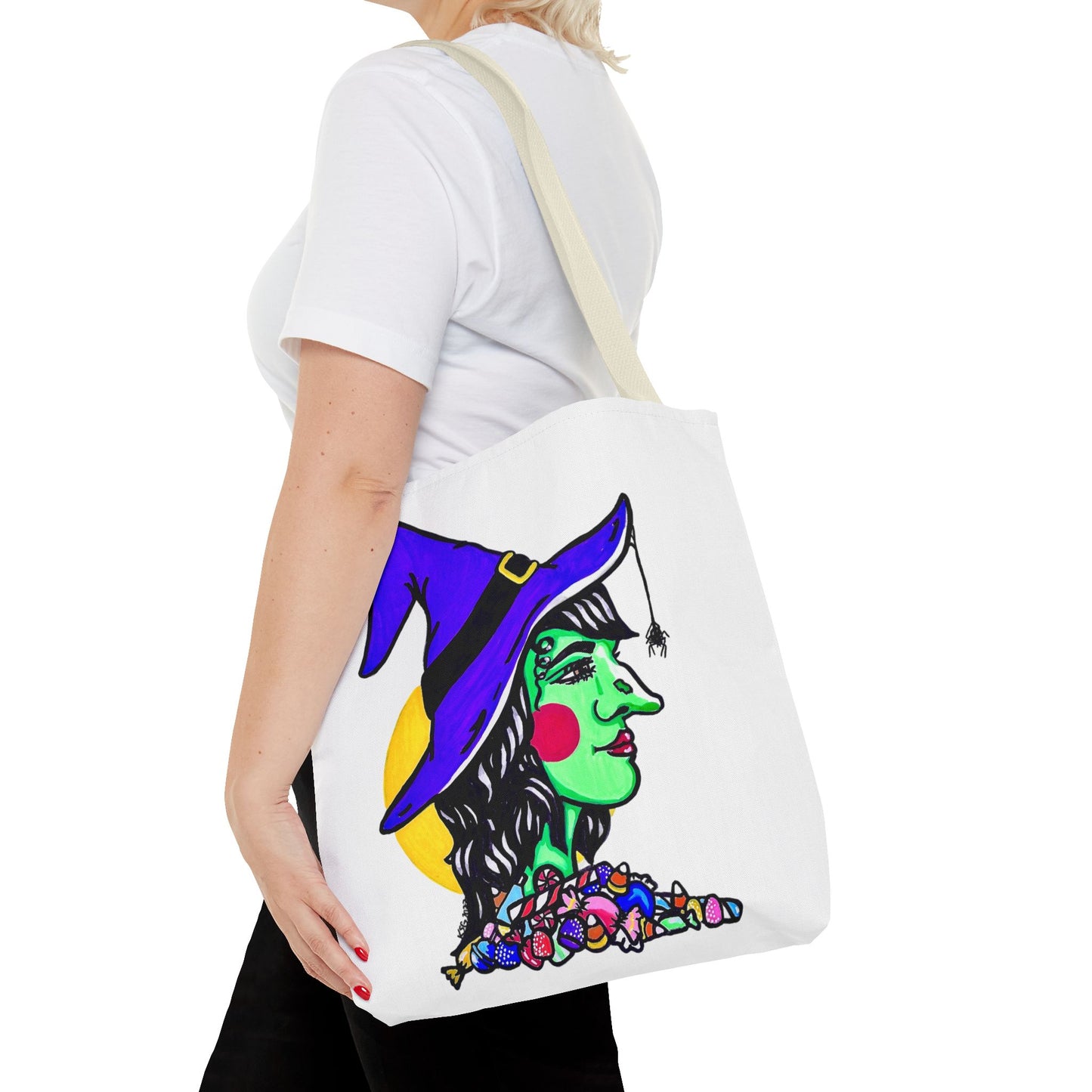 This is the Beige 16" x 16" Witch Tote Bag by Storm Garden Studio. It shows the side profile image of a witch. She is facing the right side. She has a purple witch hat with a black belt, yellow buckle, a spider crawling down the front, red rosy cheeks, red lips and assorted candy under the neck. Behind her is a bright yellow circle past her hair. It is over the shoulder of a short blonde haired woman wearing a white tea and black pants. Her hand with red nails is on the tote and she is facing the left. 