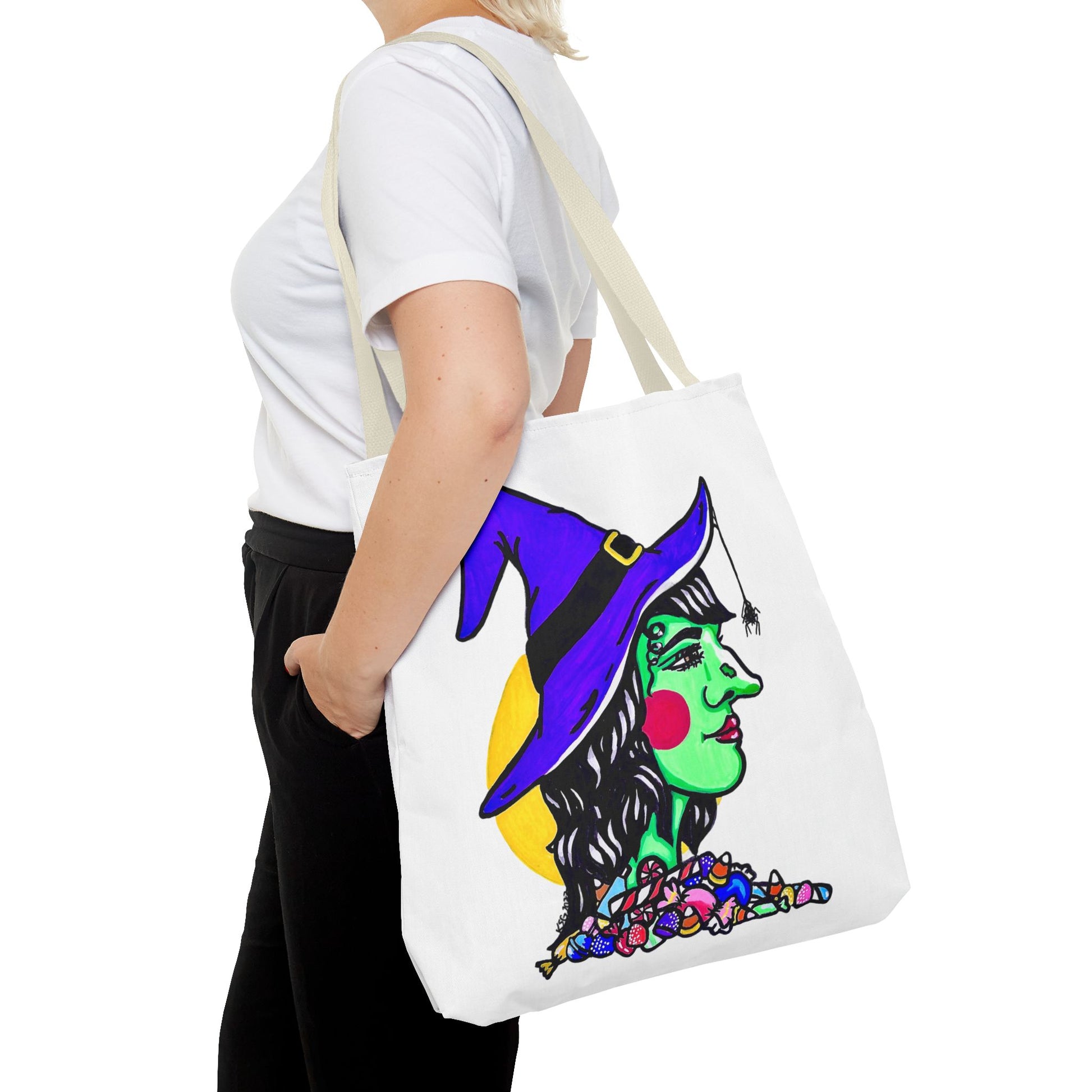 This is the Beige 18" x 18" Witch Tote Bag by Storm Garden Studio. It shows the side profile image of a witch. She is facing the right side. She has a purple witch hat with a black belt, yellow buckle, a spider crawling down the front, red rosy cheeks, red lips and assorted candy under the neck. Behind her is a bright yellow circle past her hair. It is over the shoulder of a short blonde haired woman wearing a white tea and black pants. Her hand in the pants pocket and she is facing the left. 