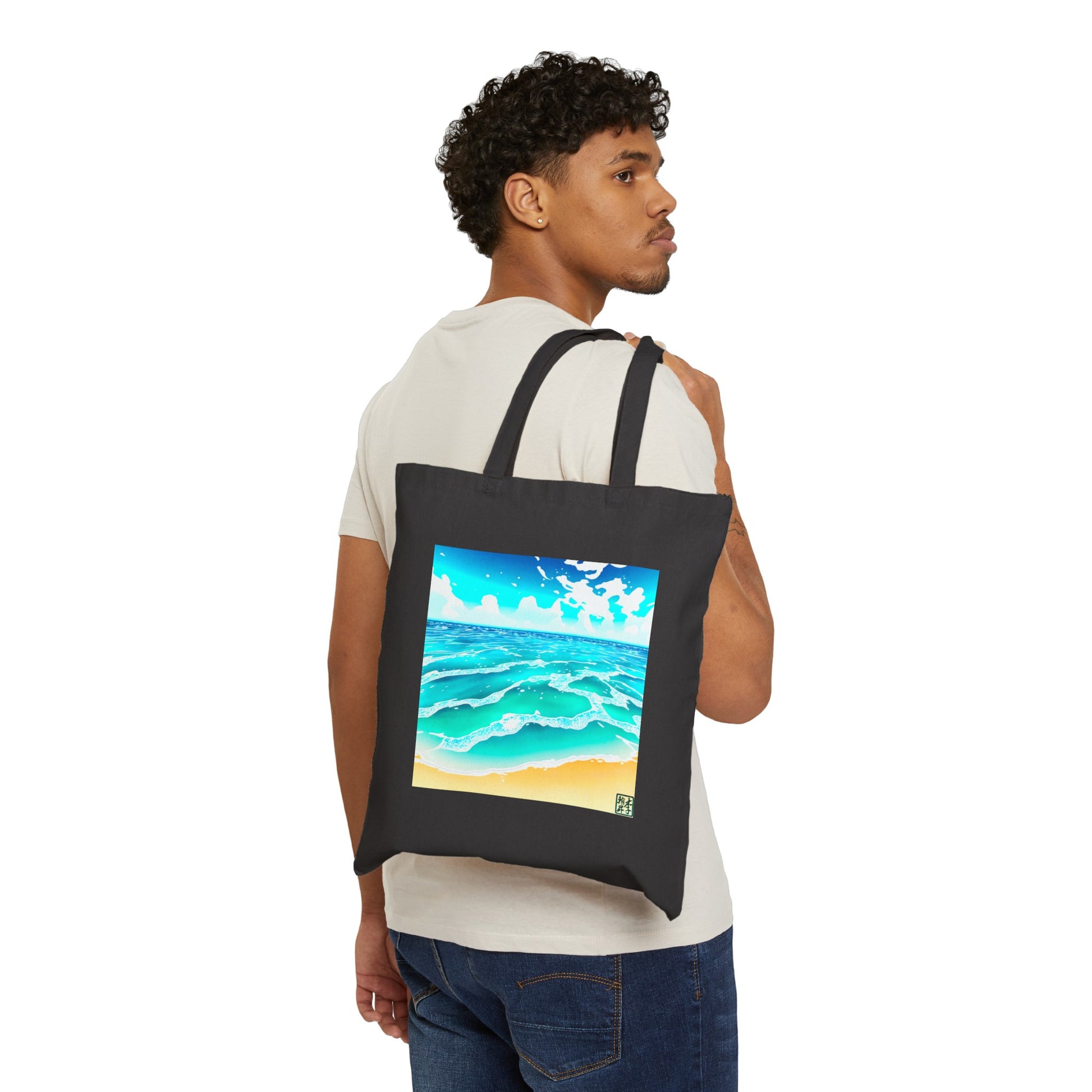 This is the Black 15" x 16" Beach Life Canvas Tote by Lee Hansheng Studios. The illustration is squared,the foreground has sand and the studio logo on the right corner outlined in blue. The waves are crashing into the sand with fluffly clouds in the back. There are mixes of blue, light blue, white, orange and tan colors. A man looking to the right is holding the handles with the tote over his shoulder showing the front and wearing a white tee and dark jeans. 