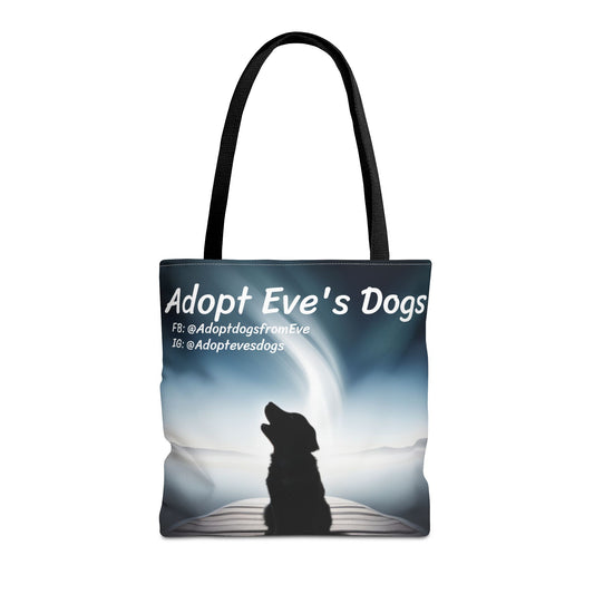 This is the Black 16" x 16" Adopt Eve's Dogs Tote with color matched handles. This is an all around print tote with "Adopt Eve's Dogs" and socials on the top in white. The print is of a shadow of a golden retriever looking to the left on a pier towards water with mountains surrounding it. The tote is against a white background.