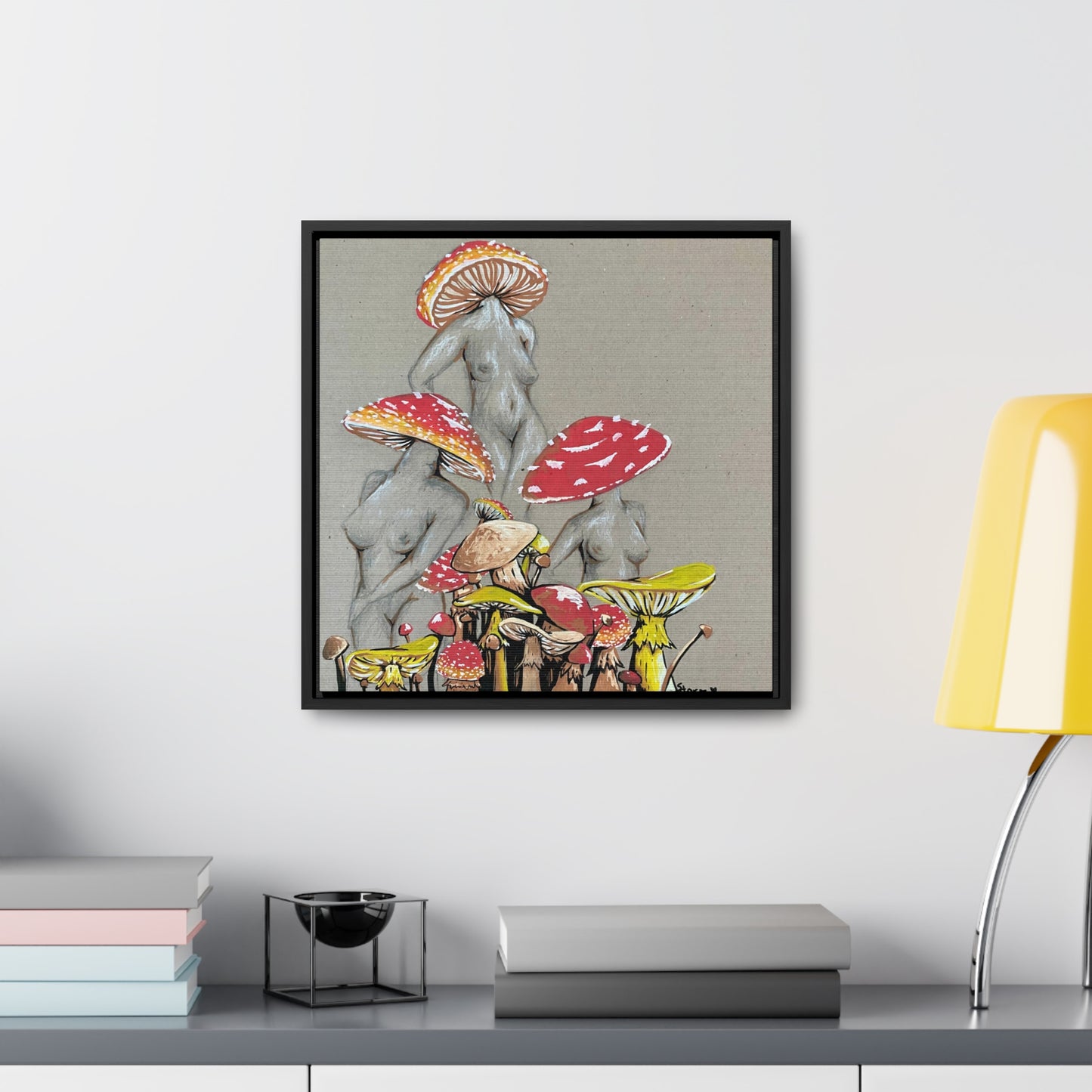 This is the 16" x 16" Miss Amanita Gallery Canvas Wrap with Black Square Frame by Storm Garden Studio. The depth of the frame is 1.25". This is an illustration of three mushroom headed nude women. The foreground has assorted mushrooms in different sizes. The three women are positioned in a triangle shape. The colors range from yellow, orange and red. The illustration is framed on a white wall above a gray stand. Yellow lamp, stack of books and decor are spread around the surface.