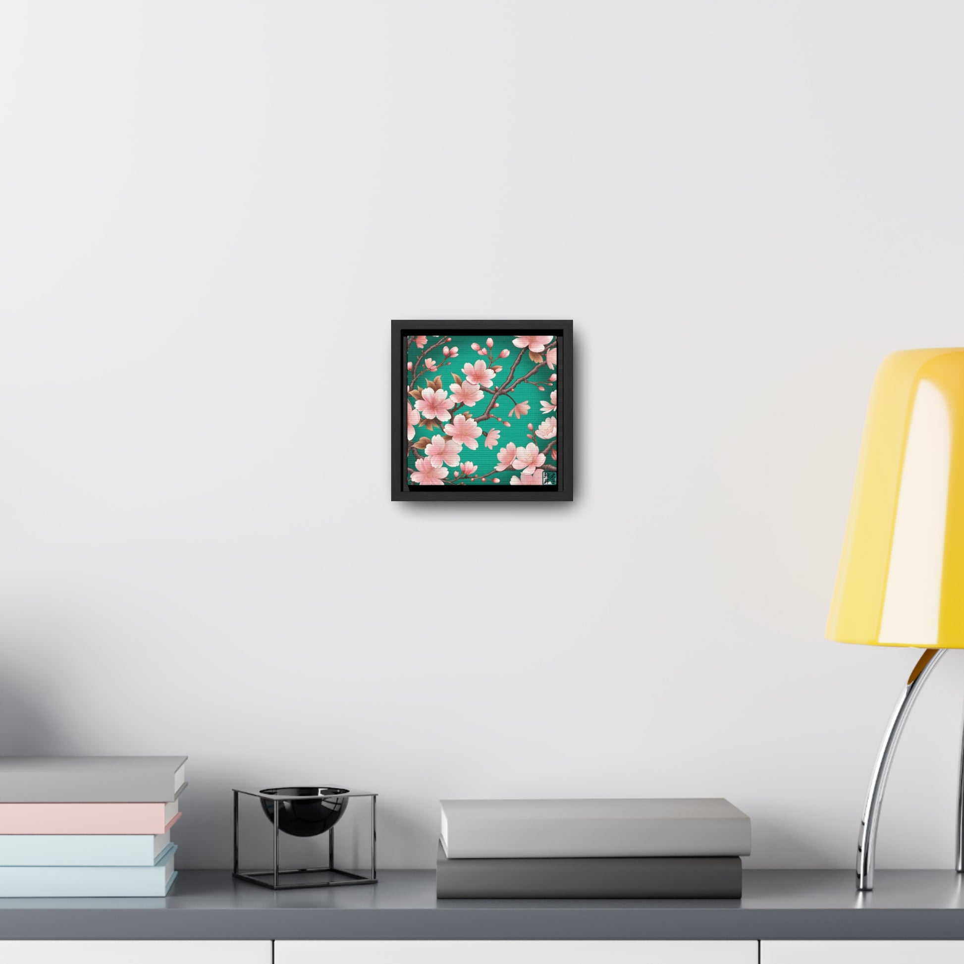 This is the Black 6" x 6" Sakura Gallery Canvas Wrap by Lee Hansheng Studios. The illustration has a blue/teal background, 3-4 thin dark wood branches with pink/light pink buds on each stem. There are blooming and already flowered sakuras on each branch. The studio's logo is on the bottom right printed in black outlined in blue. Below the canvas is a gray/white shelf, yellow lamp, a decoration, gray and pastel books on the shelf spread across it. The canvas is on a white wall.