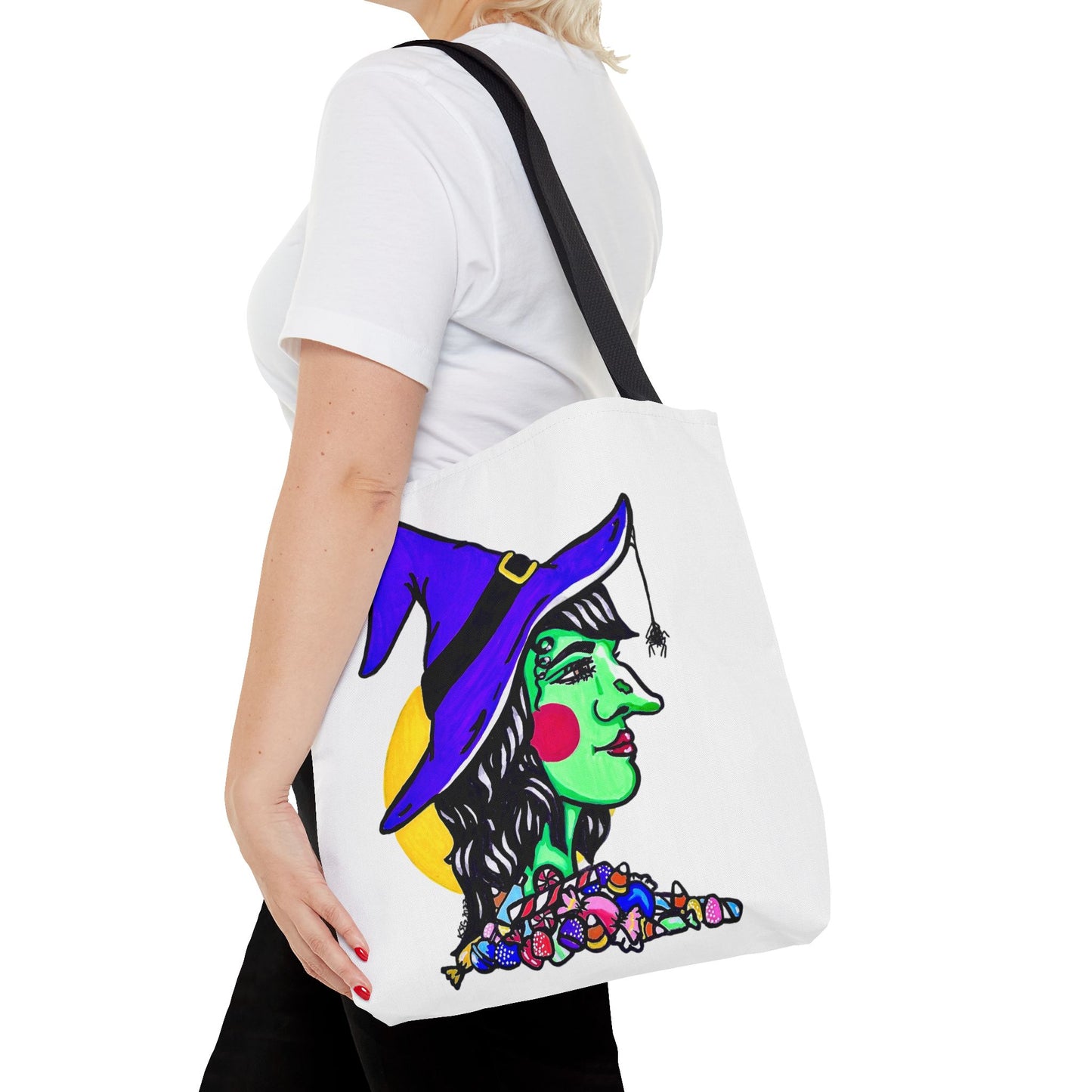 This is the Black 16" x 16" Witch Tote Bag by Storm Garden Studio. It shows the side profile image of a witch. She is facing the right side. She has a purple witch hat with a black belt, yellow buckle, a spider crawling down the front, red rosy cheeks, red lips and assorted candy under the neck. Behind her is a bright yellow circle past her hair. It is over the shoulder of a short blonde haired woman wearing a white tea and black pants. Her hand with red nails is on the tote and she is facing the left. 