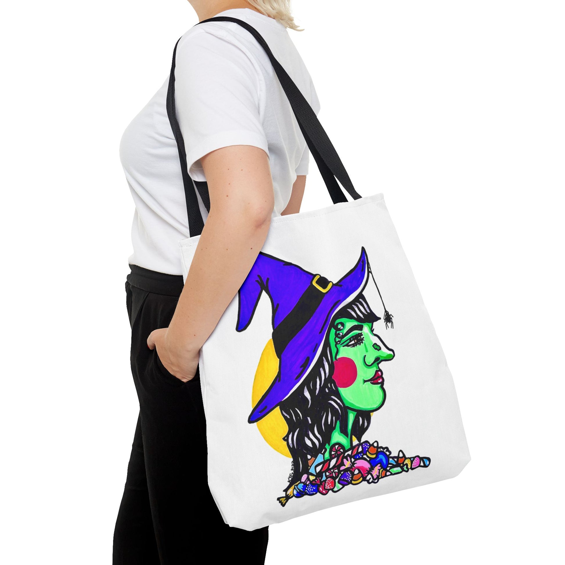 This is the Black 18" x 18" Witch Tote Bag by Storm Garden Studio. It shows the side profile image of a witch. She is facing the right side. She has a purple witch hat with a black belt, yellow buckle, a spider crawling down the front, red rosy cheeks, red lips and assorted candy under the neck. Behind her is a bright yellow circle past her hair. It is over the shoulder of a short blonde haired woman wearing a white tea and black pants. Her hand with red nails is on the tote and she is facing the left. 