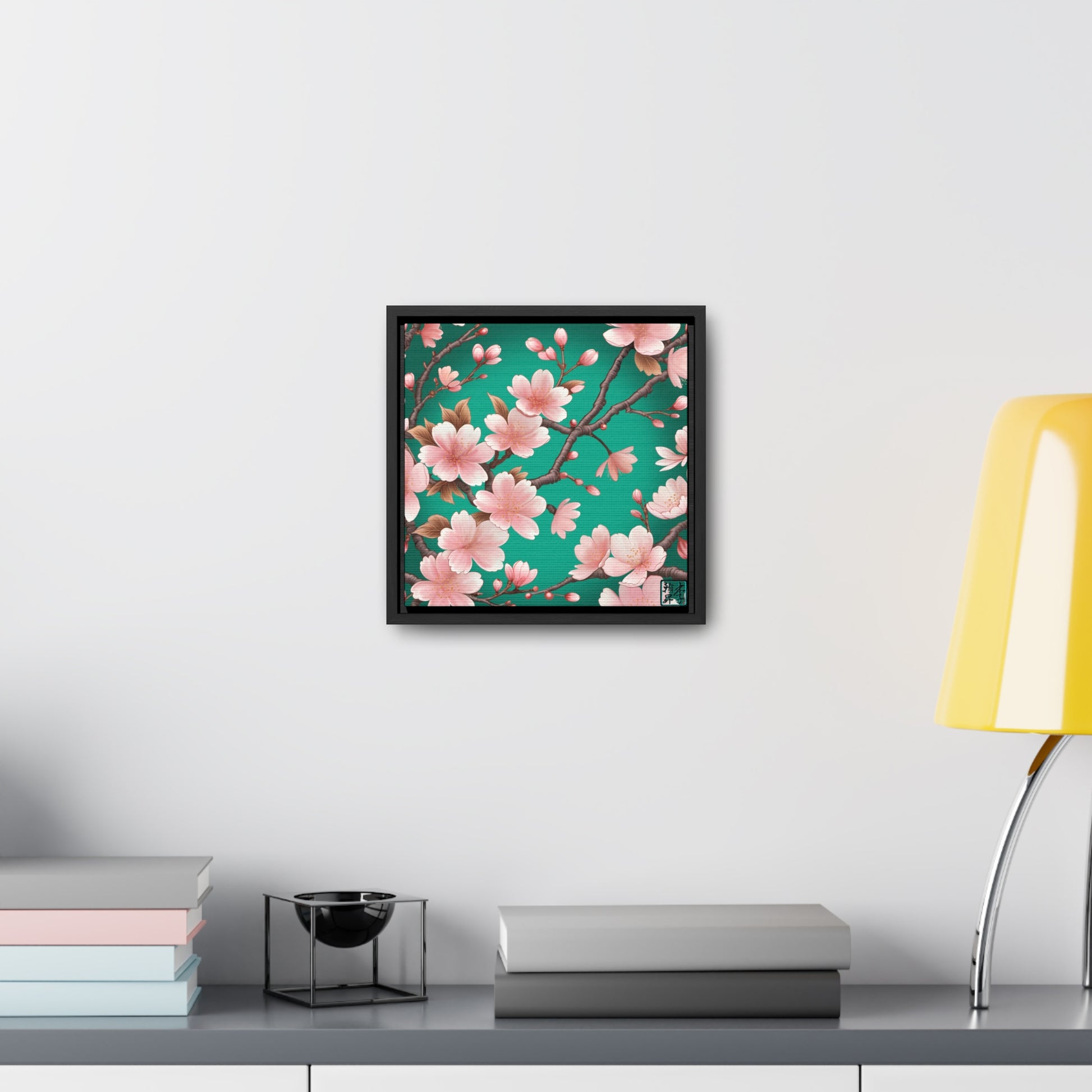 This is the Black 12" x 12" Sakura Gallery Canvas Wrap by Lee Hansheng Studios. The illustration has a blue/teal background, 3-4 thin dark wood branches with pink/light pink buds on each stem. There are blooming and already flowered sakuras on each branch. The studio's logo is on the bottom right printed in black outlined in blue. Below the canvas is a gray/white shelf, yellow lamp, a decoration, gray and pastel books on the shelf spread across it. The canvas is on a white wall.