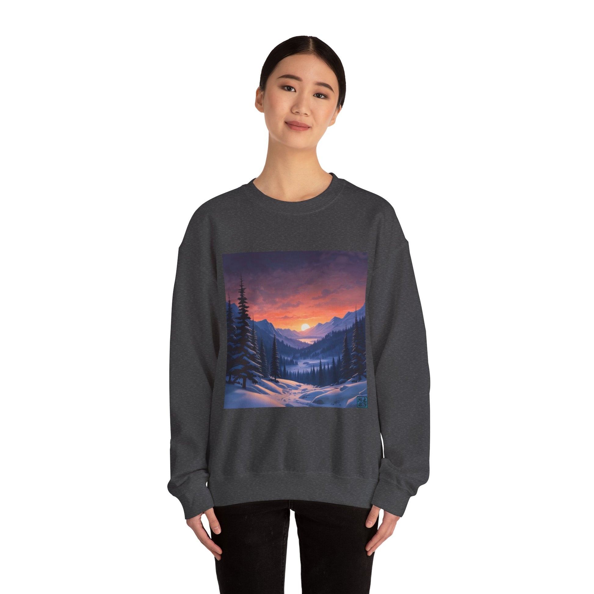 This is the Dark Heather Snow Valley Sweatshirt that is a crewneck coming in S - 5XL sizes. It is worn by an Asian lady who has black hair tied in a bun, black pants and is standing against a white background with a smile. This design is by Lee Hansheng Studios and showcases a snowy hill/mountain area with large pine trees on both sides continuing into the horizon. The sun is rising past the horizon and consist of yellow,orange,red hues which is illuminated onto the clouds,snowy field, mountains and trees. 