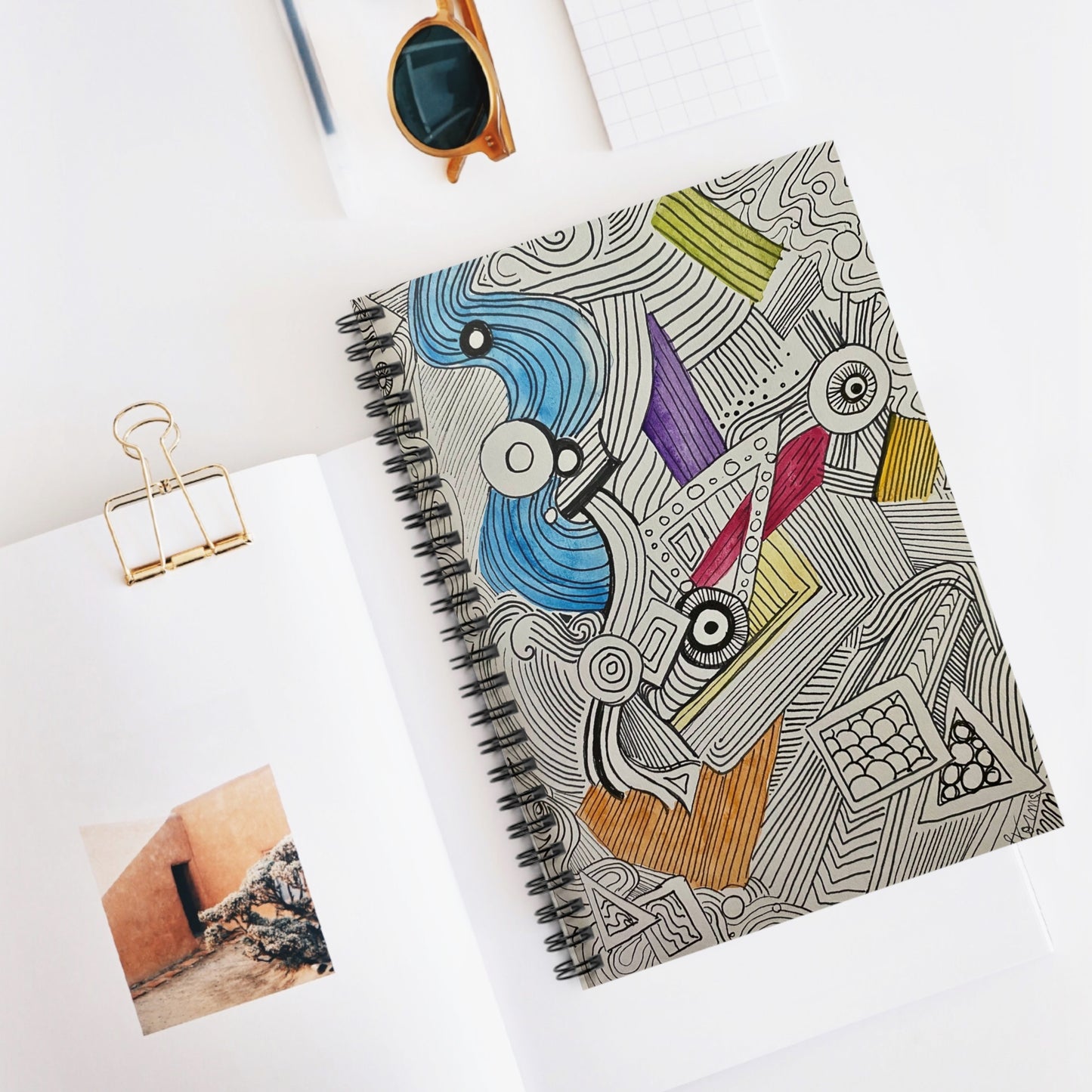 This is the Doodle Spiral Notebook with Ruled Line Paper and metal spiral binding by Storm Garden Studio. This is an image of the notebook from the front slightly to the left. The illustration is different shapes, lines and colors going in all directions. Most of it is black and white with sparse colors. There is blue, yellow, purple, red and orange. Everything is on a white surface, to the left is a gold binder clip with papers, sunglasses above the notebook and a clear black pen next to it.