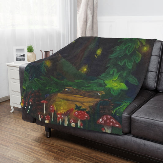 This is the 30" x 40" Forest Toad Microfiber Blanket by Storm Garden Studio. The blanket illustration is deep in the forest with different sizes of red spotted mushrooms, pinecones, vines and a toad. In the background there are fireflies, large ivy vine and a large mossy log. The blanket is spread across the left arm to the back cushion. There is dark grey leather and is paired with a white side table with a water pot, stod up picture and plant. There is a dark wood floor with white walls and sheer shades.