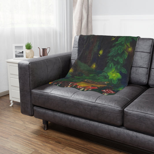 This is the 40" x 60" Forest Toad Microfiber Blanket by Storm Garden Studio. The blanket illustration is deep in the forest with different sizes of red spotted mushrooms, pinecones, vines and a toad. In the background there are fireflies, large ivy vine and a large mossy log. The blanket is spread across the left arm to the back cushion. There is dark grey leather and is paired with a white side table with a water pot, stod up picture and plant. There is a dark wood floor with white walls and sheer shades.