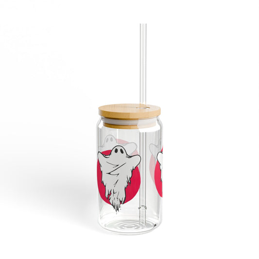 This is the 16oz Ghost Sipper Glass with lid and straw by Storm Garden Studio. This is a picture of the sipper glass slightly to the right. The ghost illustration covers the whole glass. The glass is clear with a light colored bamboo lid and a plastic straw towards the right. The ghost is white with a wrinkled and ragged cloth over it. There is a red circle behind the ghost. You can see the print throughout the glass and 1 print of the same illustration. The picture is against a white background. 