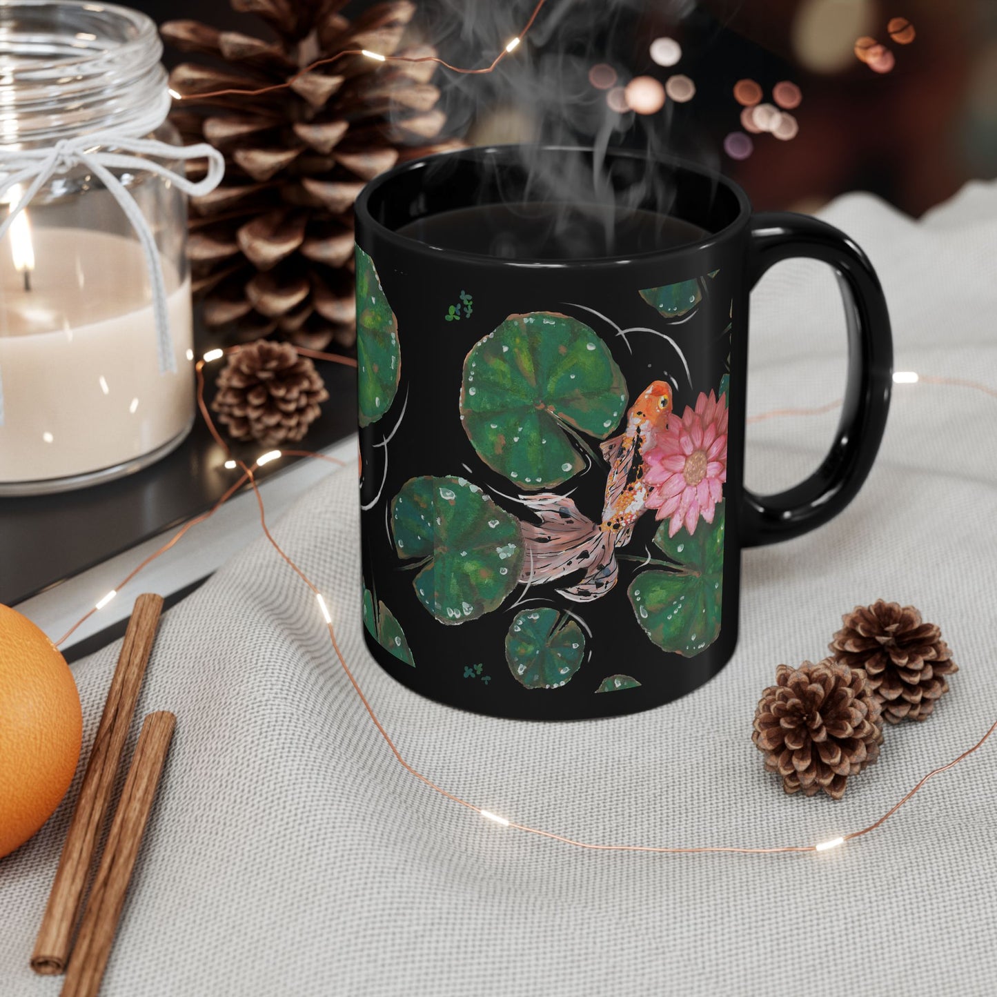 This is the 11 oz Goldfish Mug designed by Chris Foster Design. The image is shown slighty to the right. There are nine green lily pads of different sizes with water droplets reflected in clear/white. There is another speckled white,orange and black goldfish that can be fully seen swimming under a pink lily flower. The fish has three orange/pink tail fins.The water ripples and is clear matching the mug which is black.In the background there are pine cones,cinammon,orange and a candle on a surface.