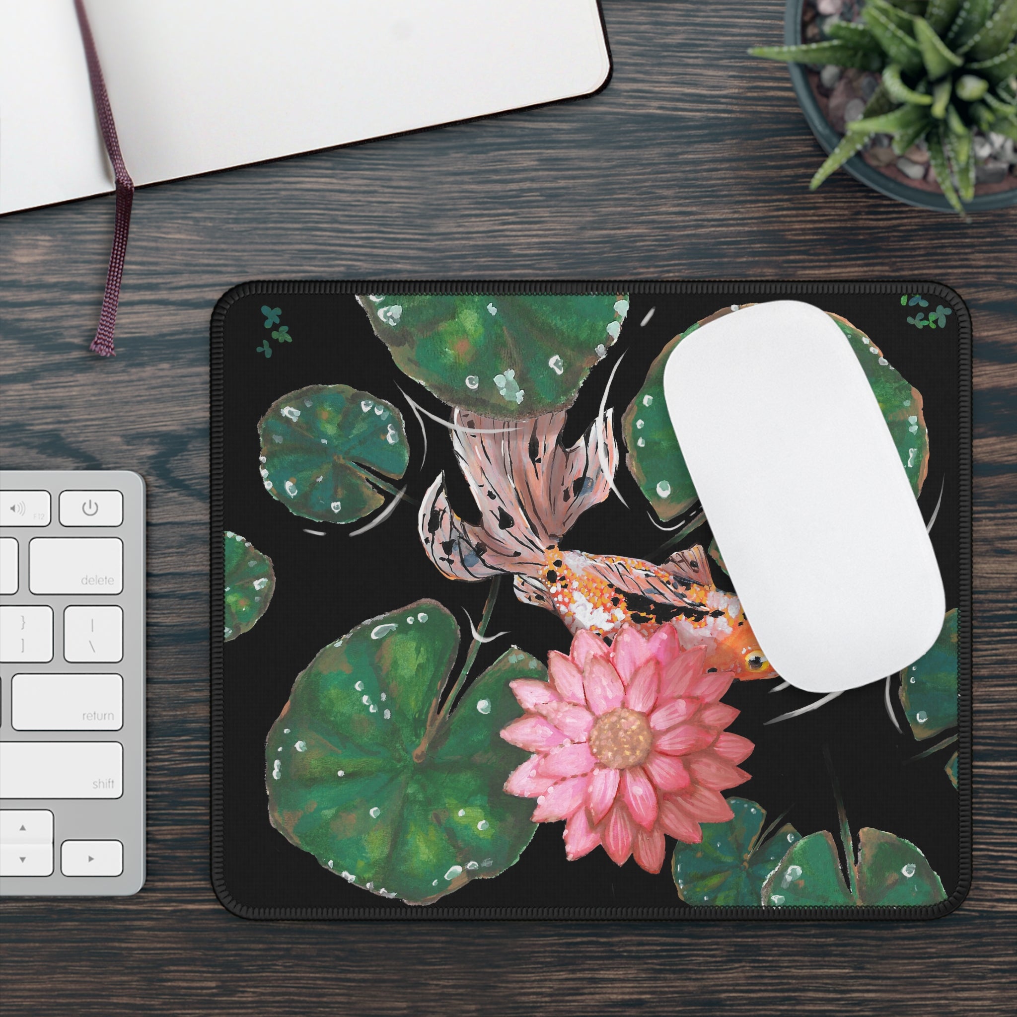 This is the Goldfish Mouse Pad by Chris Foster Design. This shows the front corner where the threading begins. The illustration has the background in black, nine dark or light green lily pads, small foliage, one orange, white and black goldfish and a pink blooming flower. The mouse pad is against a wood surface, white/silver keyboard on the left, open journal and succulent plant above it and a white mouse on the pad. 