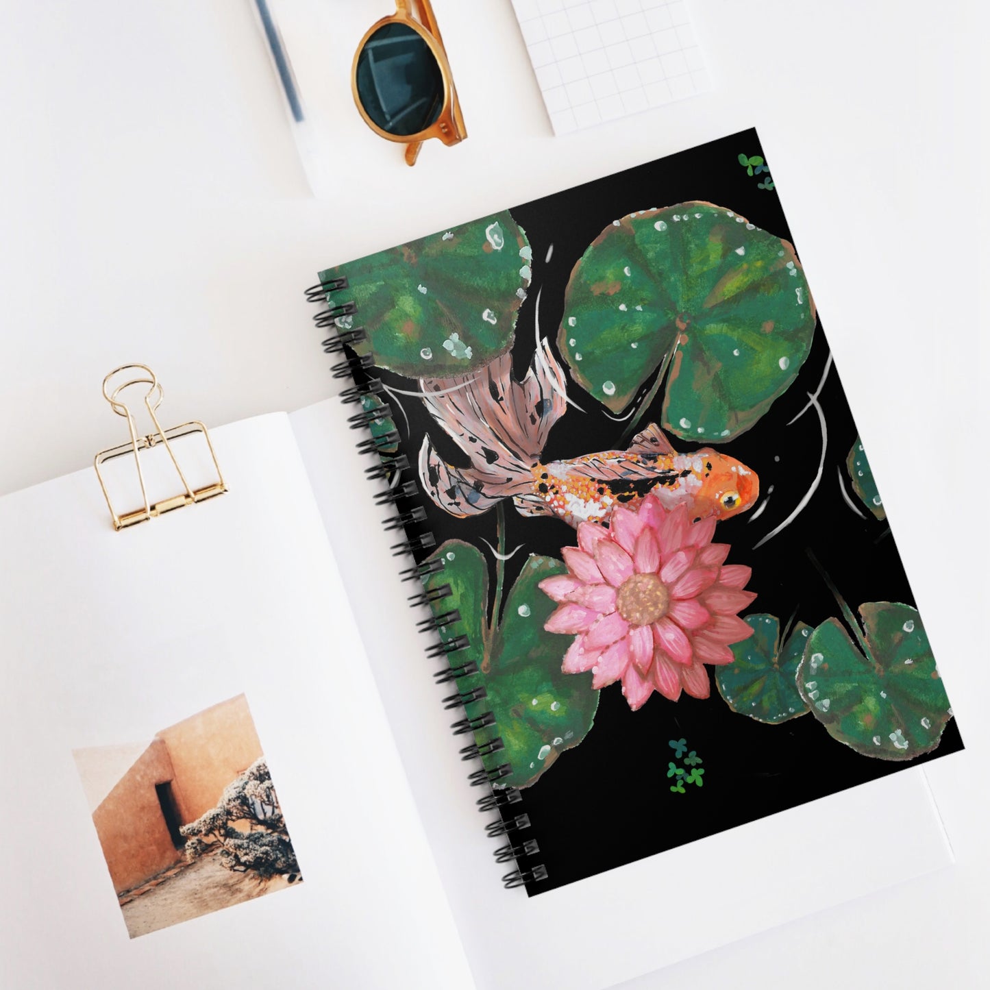 This is the Goldfish Spiral Ruled Notebook by Chris Foster Design. This shows the front of the notebook upright, slightly opened to see the black lined paper, with the illustration and the black spiral bind. The illustration is squared, the background is black, nine dark or light green lily pads, small foliage, one orange, white and black goldfish and a pink blooming flower. To the left is paper with a picture, gold binder clip, to the top is a gold/yellow sunglasses, black pen and squared paper.