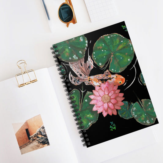 This is the Goldfish Spiral Ruled Notebook by Chris Foster Design. This shows the front of the notebook upright, slightly opened to see the black lined paper, with the illustration and the black spiral bind. The illustration is squared, the background is black, nine dark or light green lily pads, small foliage, one orange, white and black goldfish and a pink blooming flower. To the left is paper with a picture, gold binder clip, to the top is a gold/yellow sunglasses, black pen and squared paper.