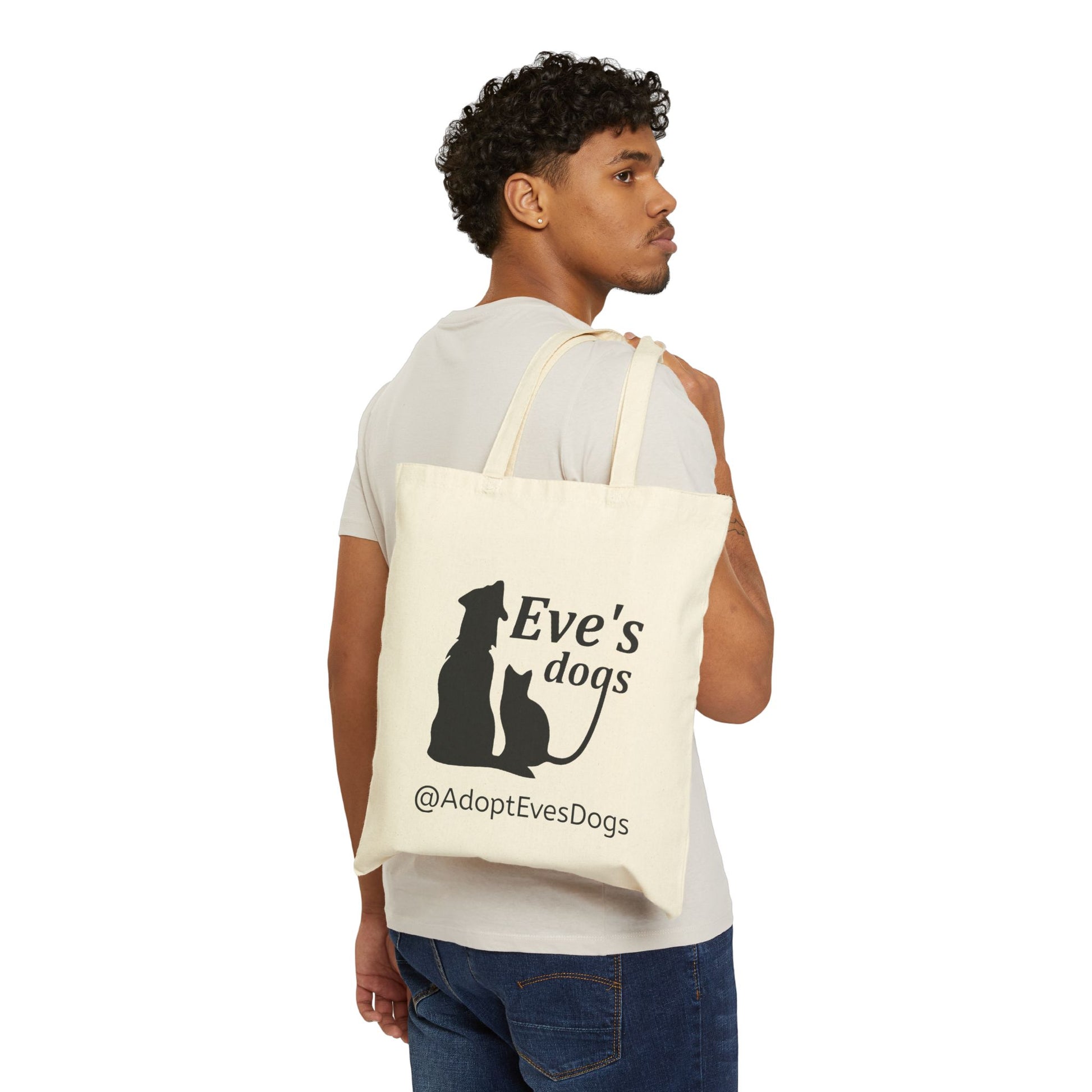 This is the Natural 15" x 16" Eve's Dogs Canvas Tote. The tote has a picture of a cat and dog in black. There is a print that says, "Eve's dogs and @AdoptEvesDogs" in black. There is an African American gentleman hanging the bag over his shoulder wearing a beige tee and jeans.