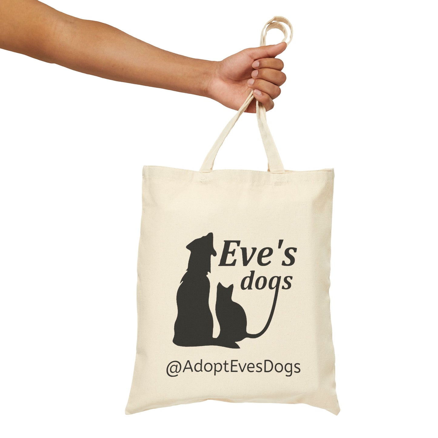 This is the Natural 15" x 16" Eve's Dogs Canvas Tote. The tote has a picture of a cat and dog in black. There is a print that says, "Eve's dogs and @AdoptEvesDogs" in black. There is an upper arm to the hand grabbing the tote from the left.