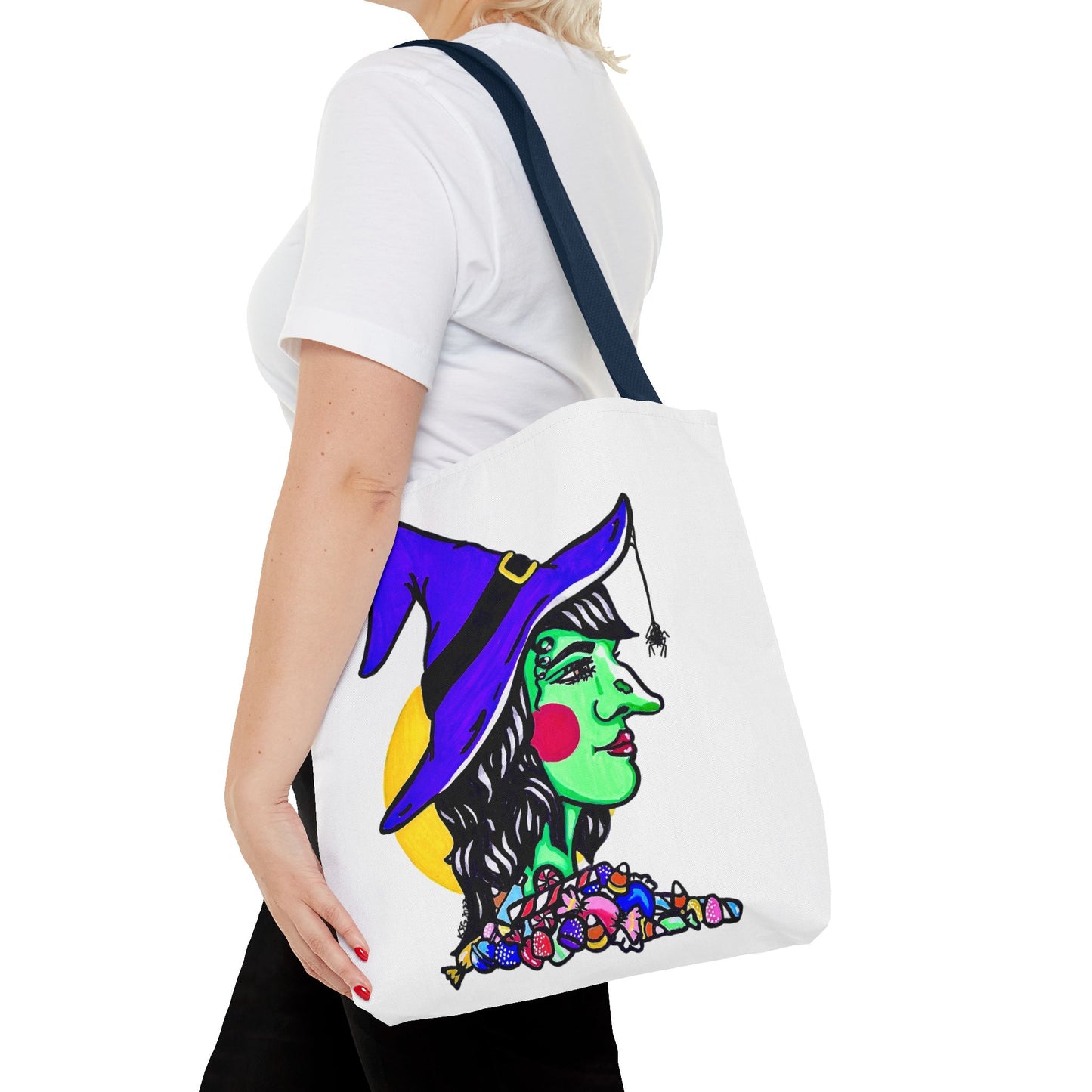 This is the Navy 16" x 16" Witch Tote Bag by Storm Garden Studio. It shows the side profile image of a witch. She is facing the right side. She has a purple witch hat with a black belt, yellow buckle, a spider crawling down the front, red rosy cheeks, red lips and assorted candy under the neck. Behind her is a bright yellow circle past her hair. It is over the shoulder of a short blonde haired woman wearing a white tea and black pants. Her hand with red nails is on the tote and she is facing the left. 