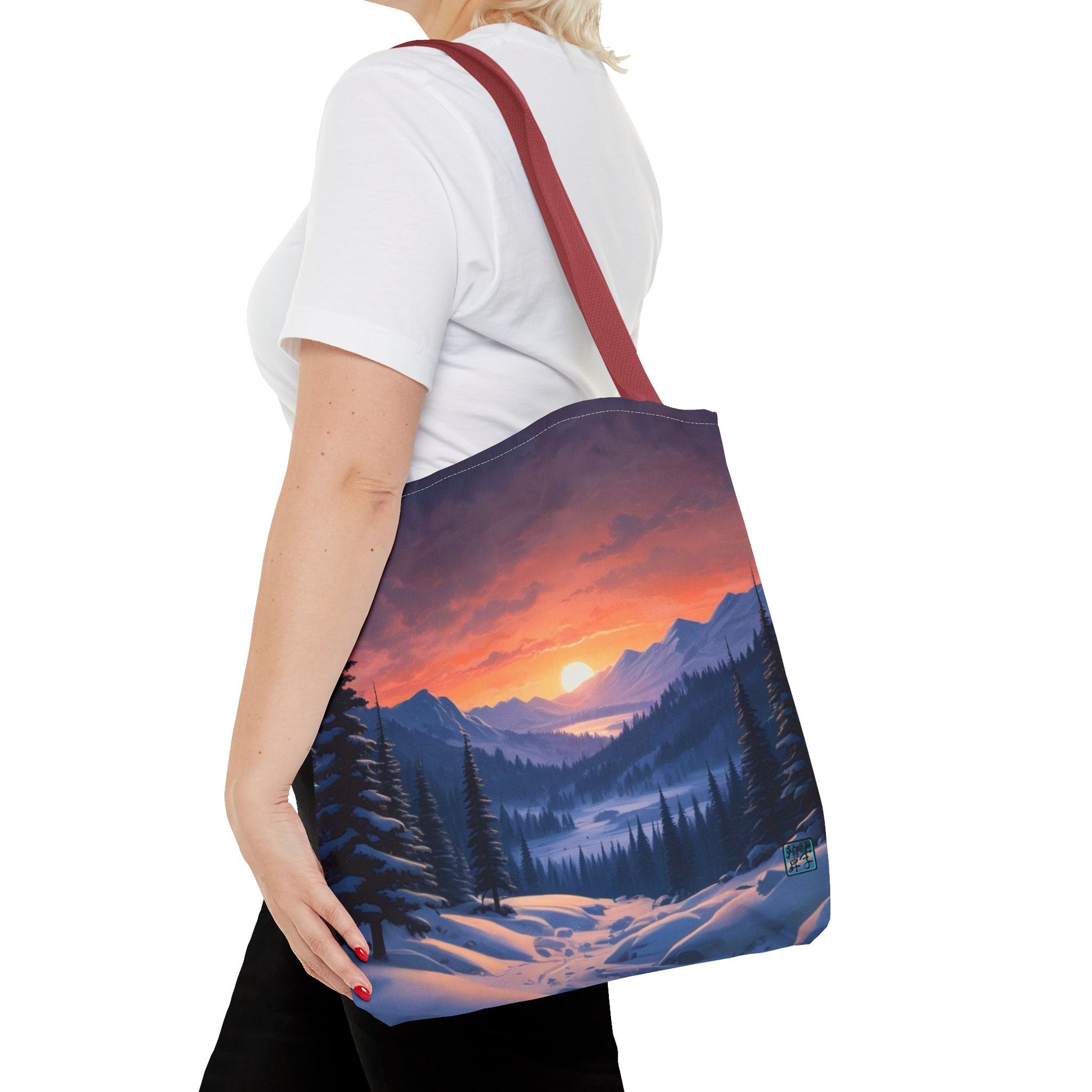 The Red 16" x 16" Snow Valley Tote Bag has color matched handles. A lady with short blonde hair,white tee plus black pants with tote around their shoulder and hand in pocket against a white background.The design by Lee Hansheng Studios showcases a snowy hill/mountain area with large pine trees on both sides continuing into the horizon. The sun rises past the horizon consisting of yellow,orange,red hues illuminating onto wispy clouds,snowy field, mountains and dark green pine trees. 