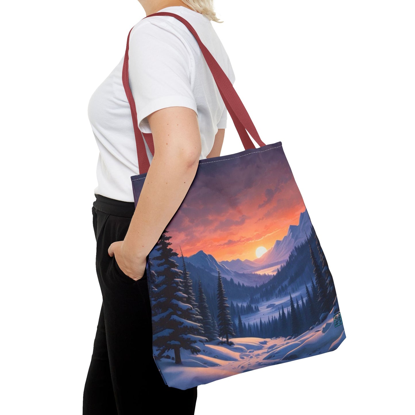 The Red 18" x 18" Snow Valley Tote Bag has color matched handles. A lady with short blonde hair,white tee plus black pants with tote around their shoulder and hand in pocket against a white background.The design by Lee Hansheng Studios showcases a snowy hill/mountain area with large pine trees on both sides continuing into the horizon. The sun rises past the horizon consisting of yellow,orange,red hues illuminating onto wispy clouds,snowy field, mountains and dark green pine trees. 