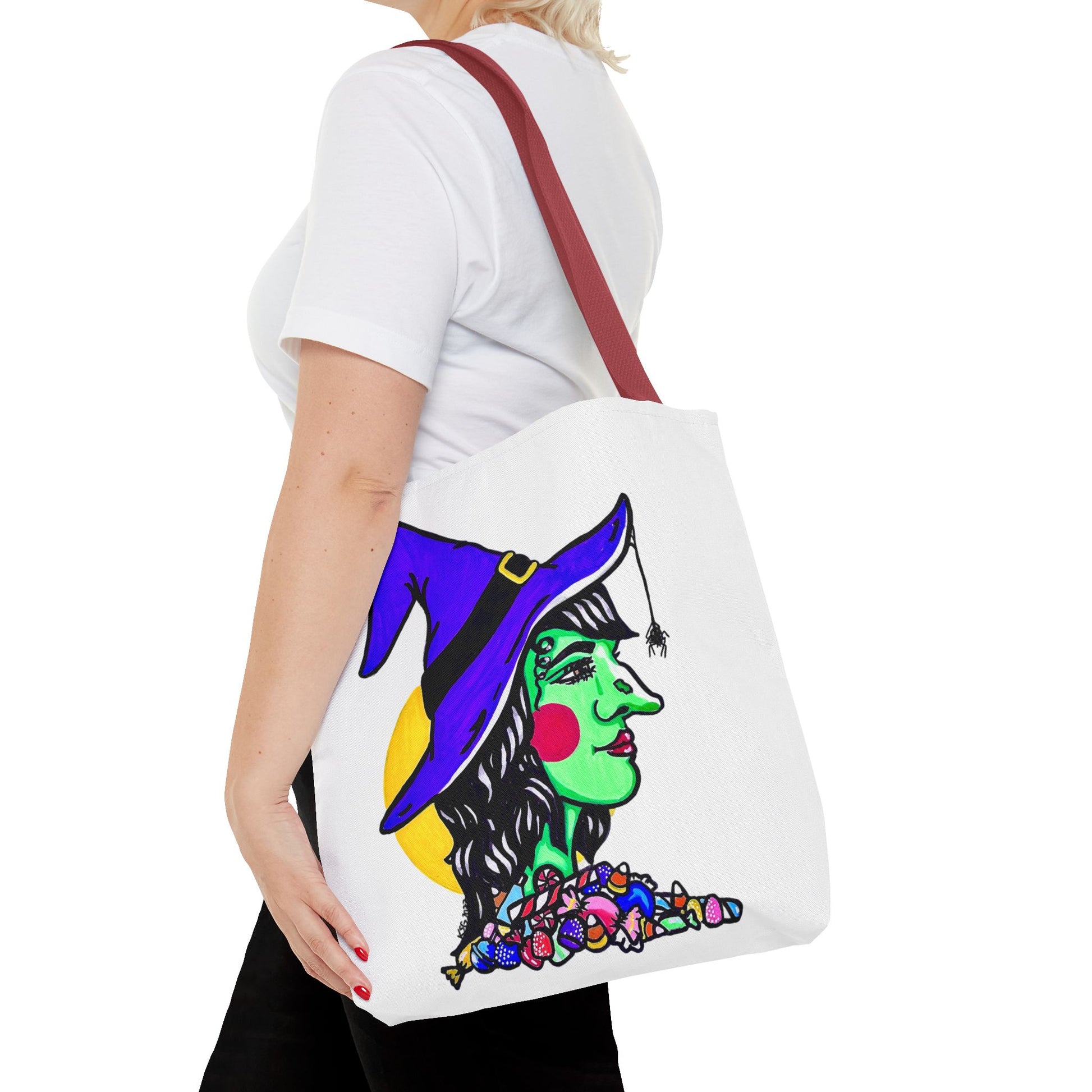 This is the Red 16" x 16" Witch Tote Bag by Storm Garden Studio. It shows the side profile image of a witch. She is facing the right side. She has a purple witch hat with a black belt, yellow buckle, a spider crawling down the front, red rosy cheeks, red lips and assorted candy under the neck. Behind her is a bright yellow circle past her hair. It is over the shoulder of a short blonde haired woman wearing a white tea and black pants. Her hand with red nails is on the tote and she is facing the left. 