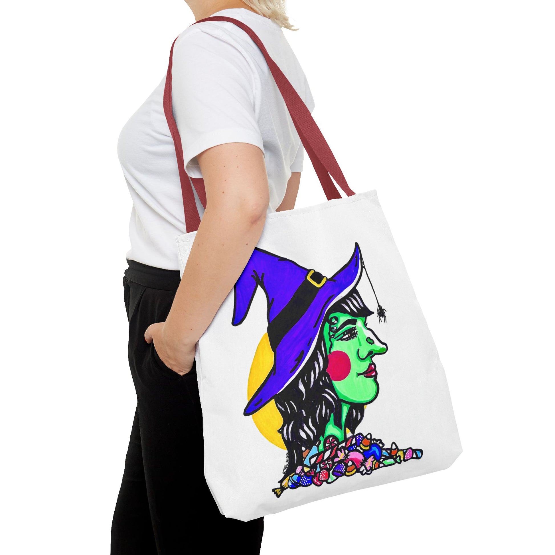 This is the Red 18" x 18" Witch Tote Bag by Storm Garden Studio. It shows the side profile image of a witch. She is facing the right side. She has a purple witch hat with a black belt, yellow buckle, a spider crawling down the front, red rosy cheeks, red lips and assorted candy under the neck. Behind her is a bright yellow circle past her hair. It is over the shoulder of a short blonde haired woman wearing a white tea and black pants. Her hand in the pants pockte and she is facing the left. 