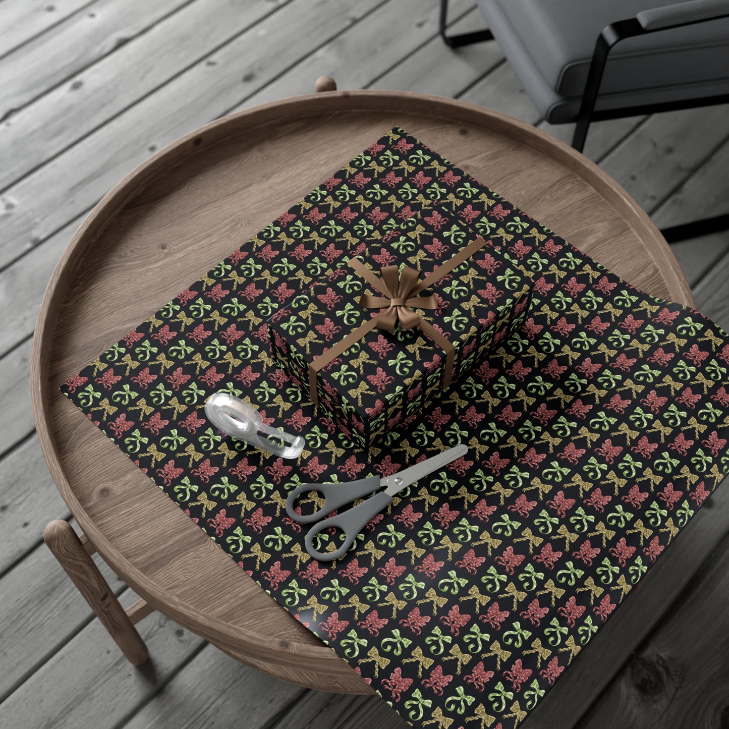 This is the 30" x 20" Matte Ribbon Gift Wrap by CFD. It is multiple illustrations of ribbons in different styles. It is placed on a small dark brown wood coffee side table. On top is the gift wrap roll on the left and below is a sheet that was cut out with a rectangular gift wrapped slightly to the right. A brown string covers each side and tied in a bow in the middle. Below is small tan scissors and clear tape roll. The background is a gray wood flooring and in the upper right is a faux leather gray chair.