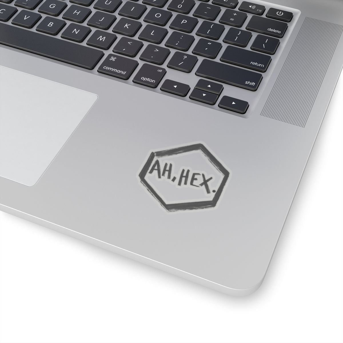 This is the Transparent 2" x 2" Ah, Hex Kiss-Cut Sticker by Tidy Tea Witch. The writing is in black and says. "AH, HEX." in the middle of the sticker. There is no illustration or image on it but is hexsagon shaped. The sticker is on a gray laptop with the black and white keyboard above and a gray/white mousepad to the left. The image is against a white background.