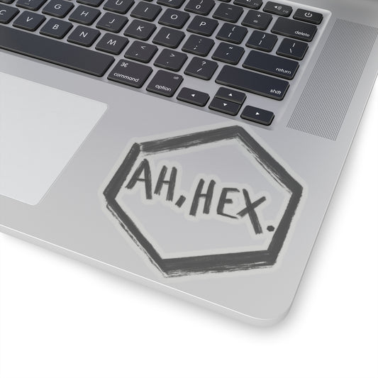 This is the Transparent 4" x 4" Ah, Hex Kiss-Cut Sticker by Tidy Tea Witch. The writing is in black and says. "AH, HEX." in the middle of the sticker. There is no illustration or image on it but is hexsagon shaped. The sticker is on a gray laptop with the black and white keyboard above and a gray/white mousepad to the left. The image is against a white background.